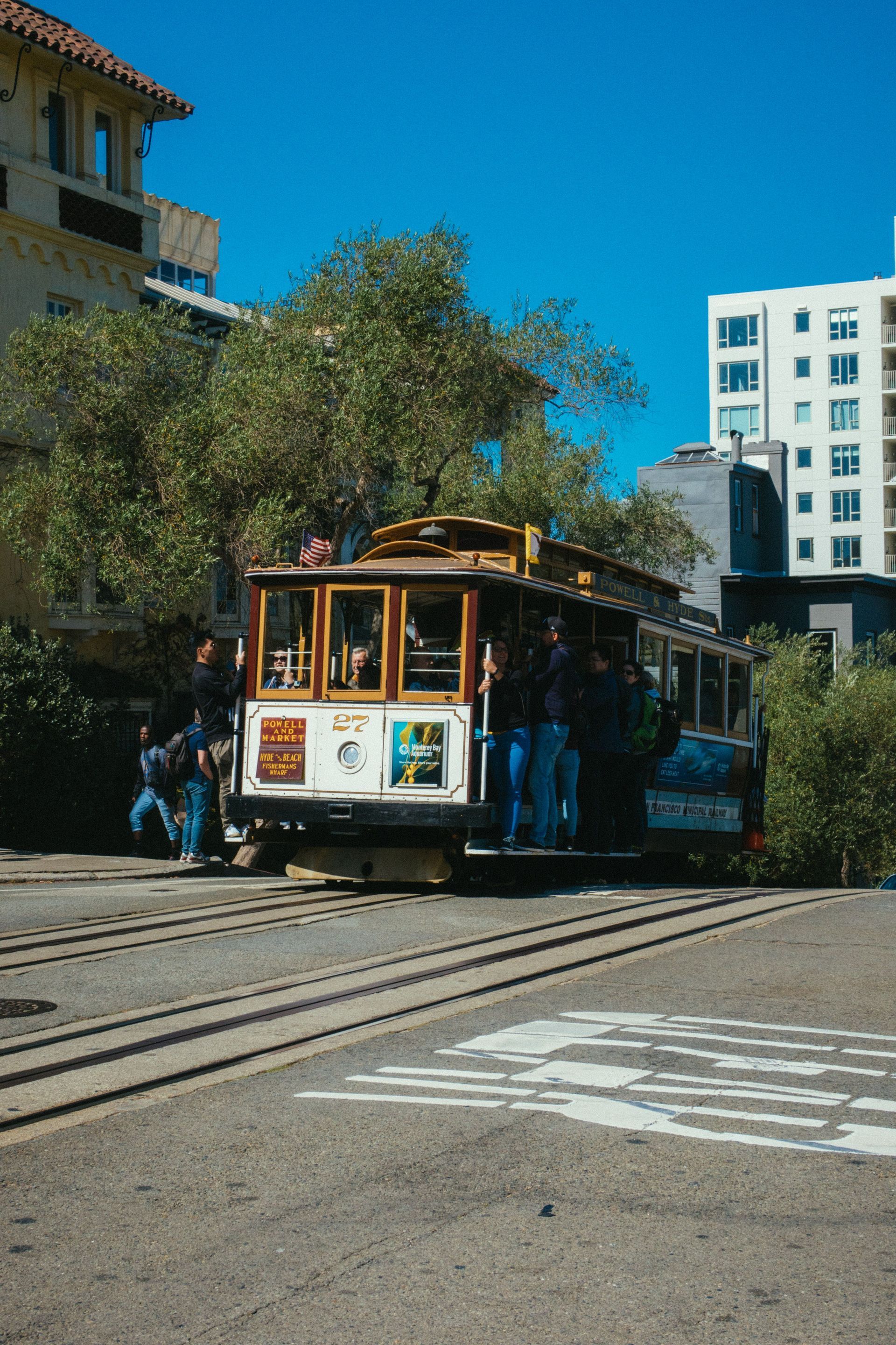 Cable car on a city street with trees and tall buildings, under a bright blue sky
