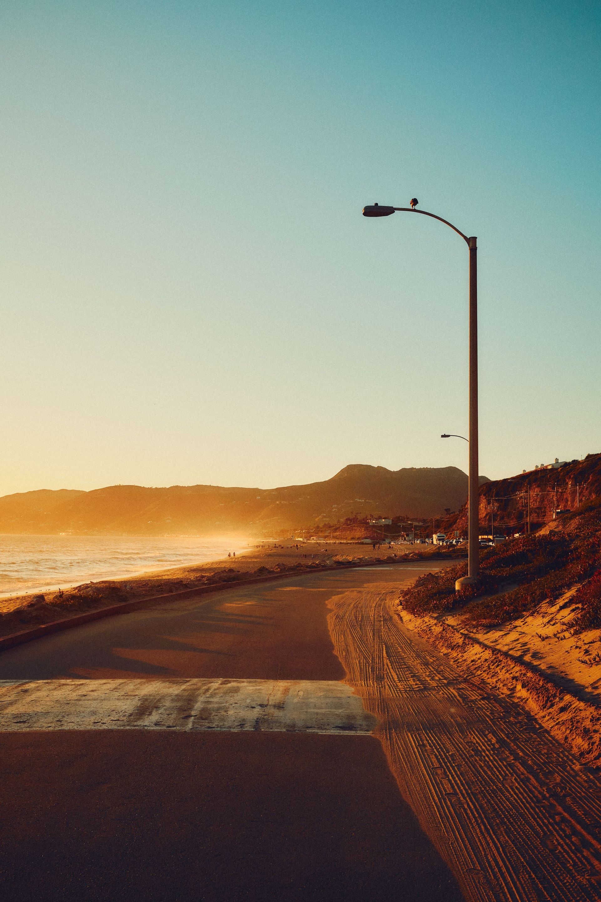 Sunset-lit coastal road curving along the shore, with a streetlamp and glowing ocean haze.
