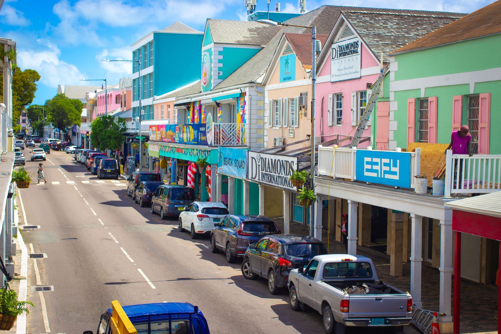 Colorful row of pastel shopfronts along a sunny street with parked cars and a clear blue sky.