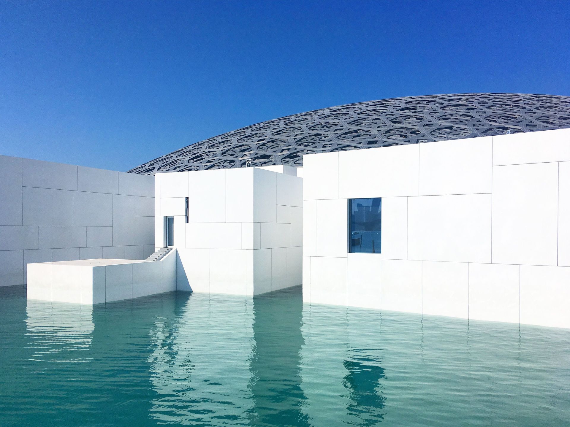 White modern building beside turquoise water under a clear blue sky, with a rocky hill in the background