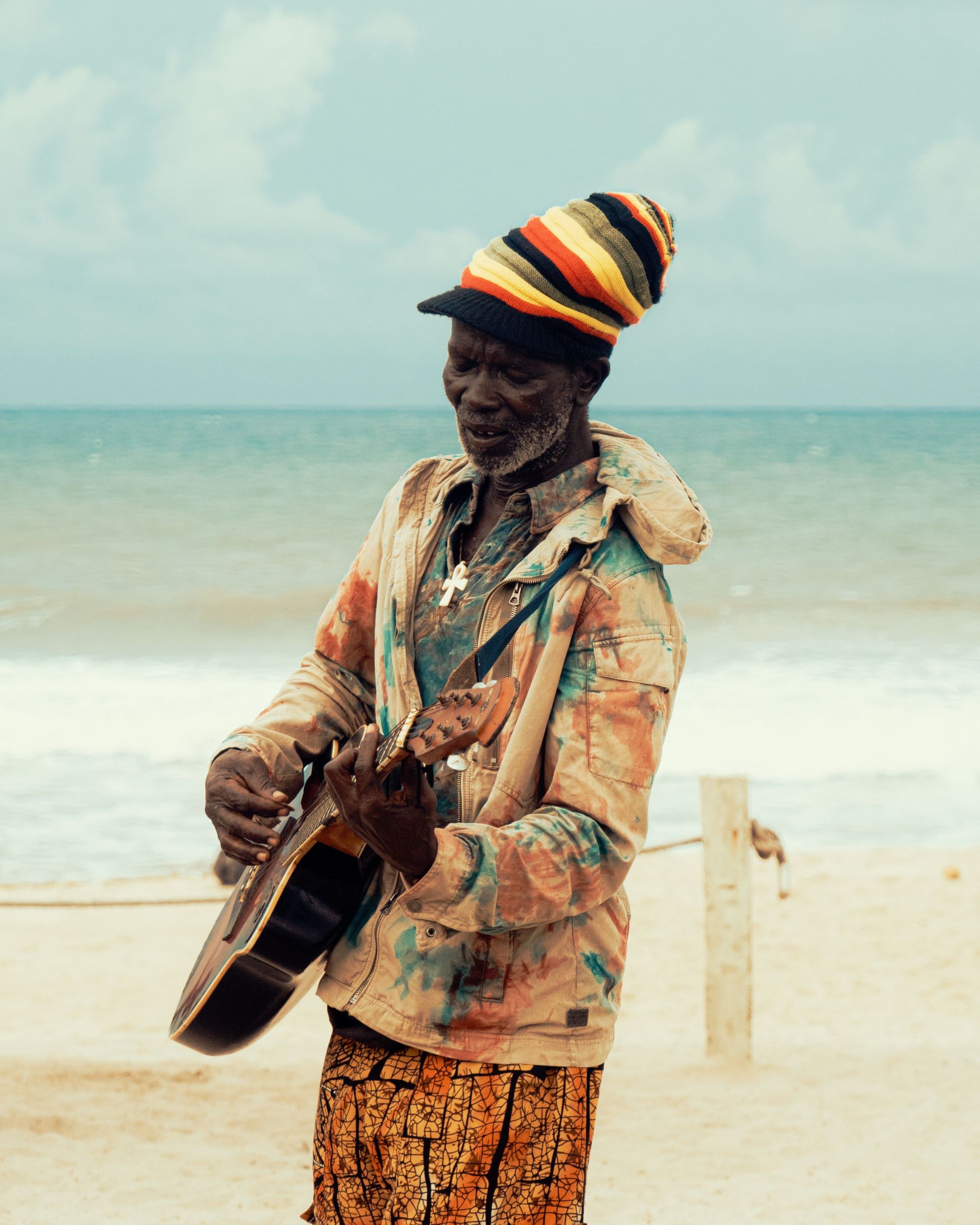 Street musician playing guitar on a beach, wearing a striped knit hat and patterned clothes.