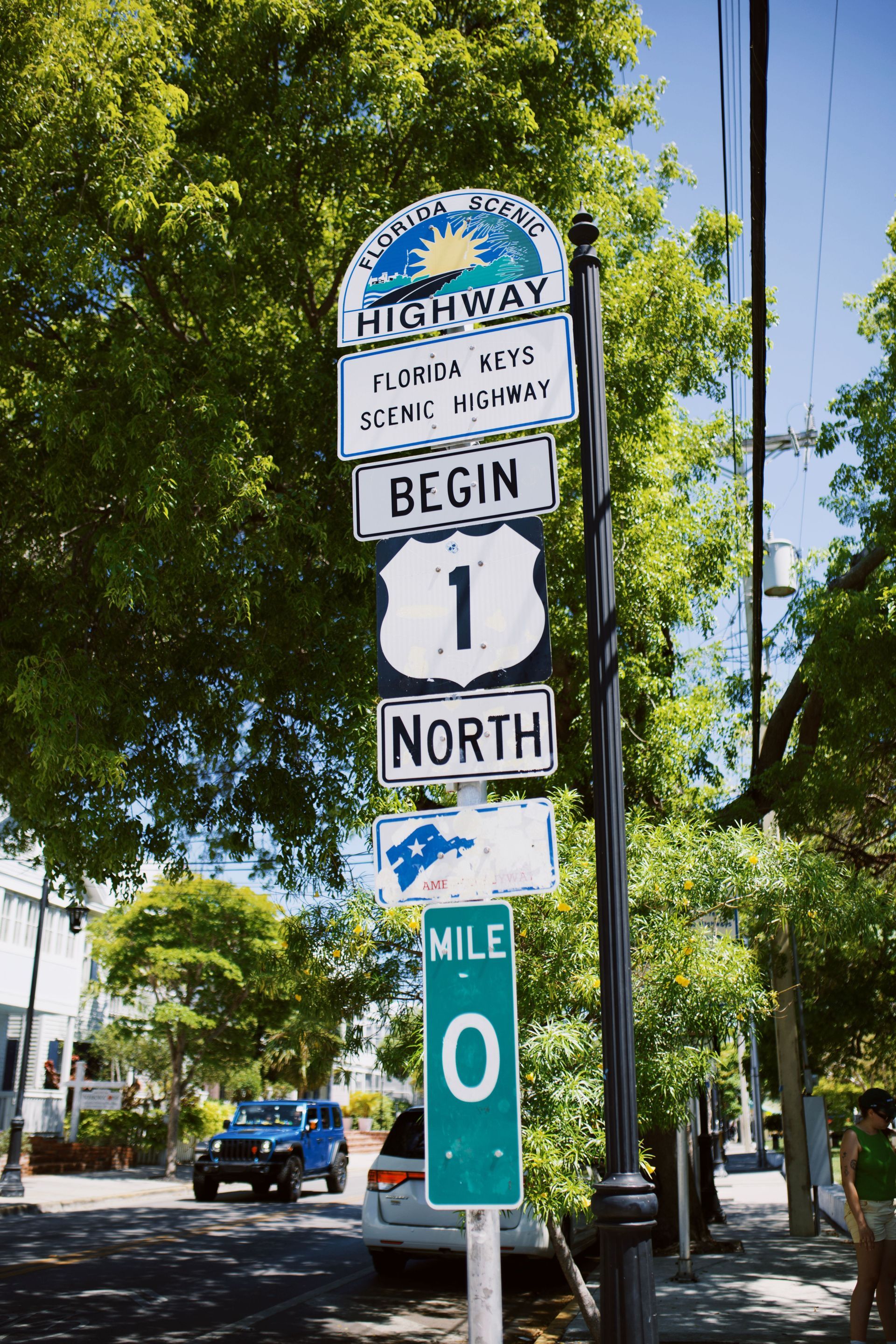 Florida Keys Highway sign: Begin 1 North, Mile 0, on a sunny street with trees and parked cars.
