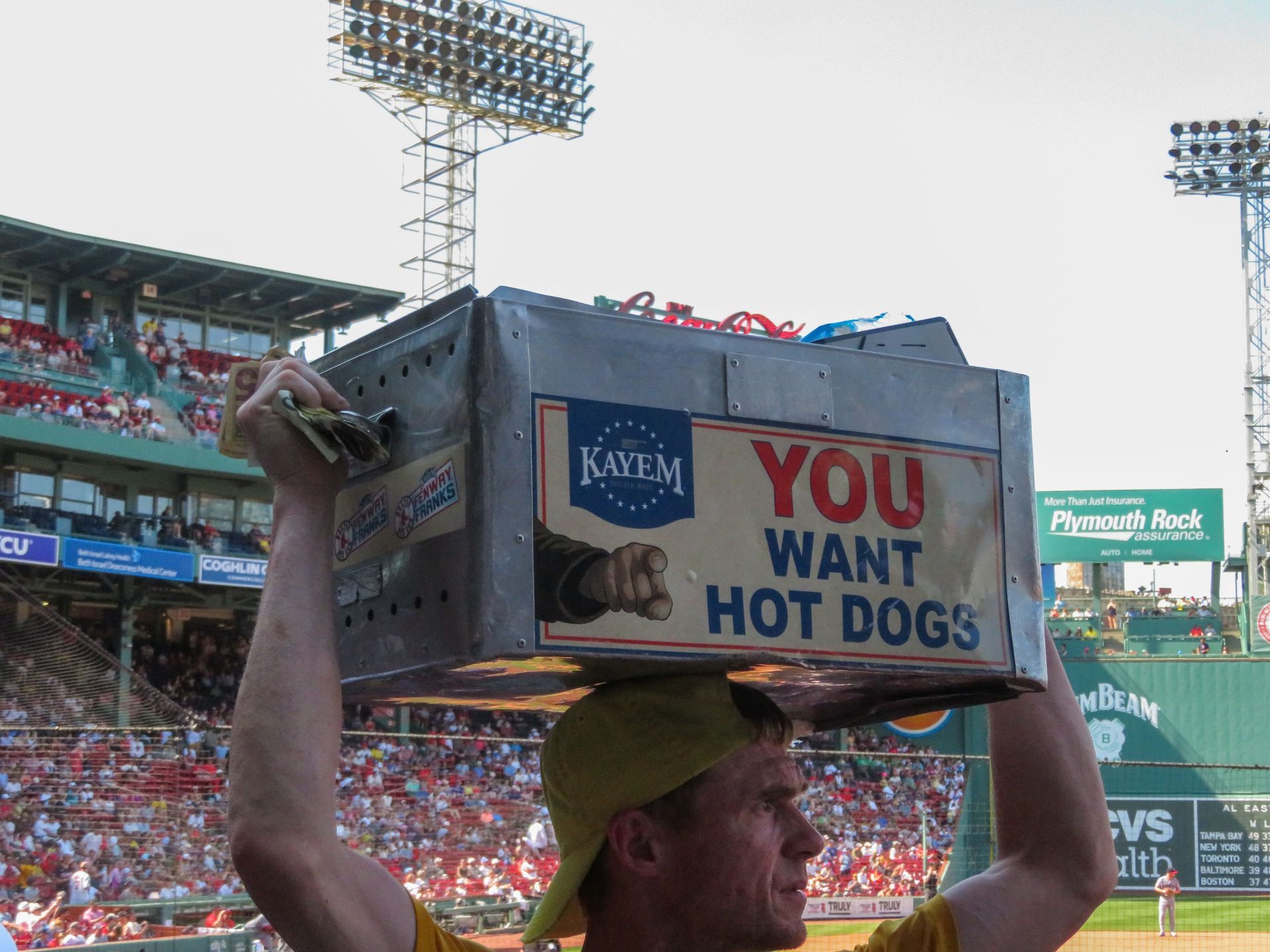 Man at a baseball stadium carries a box sign reading “YOU WANT HOT DOG” on his head.