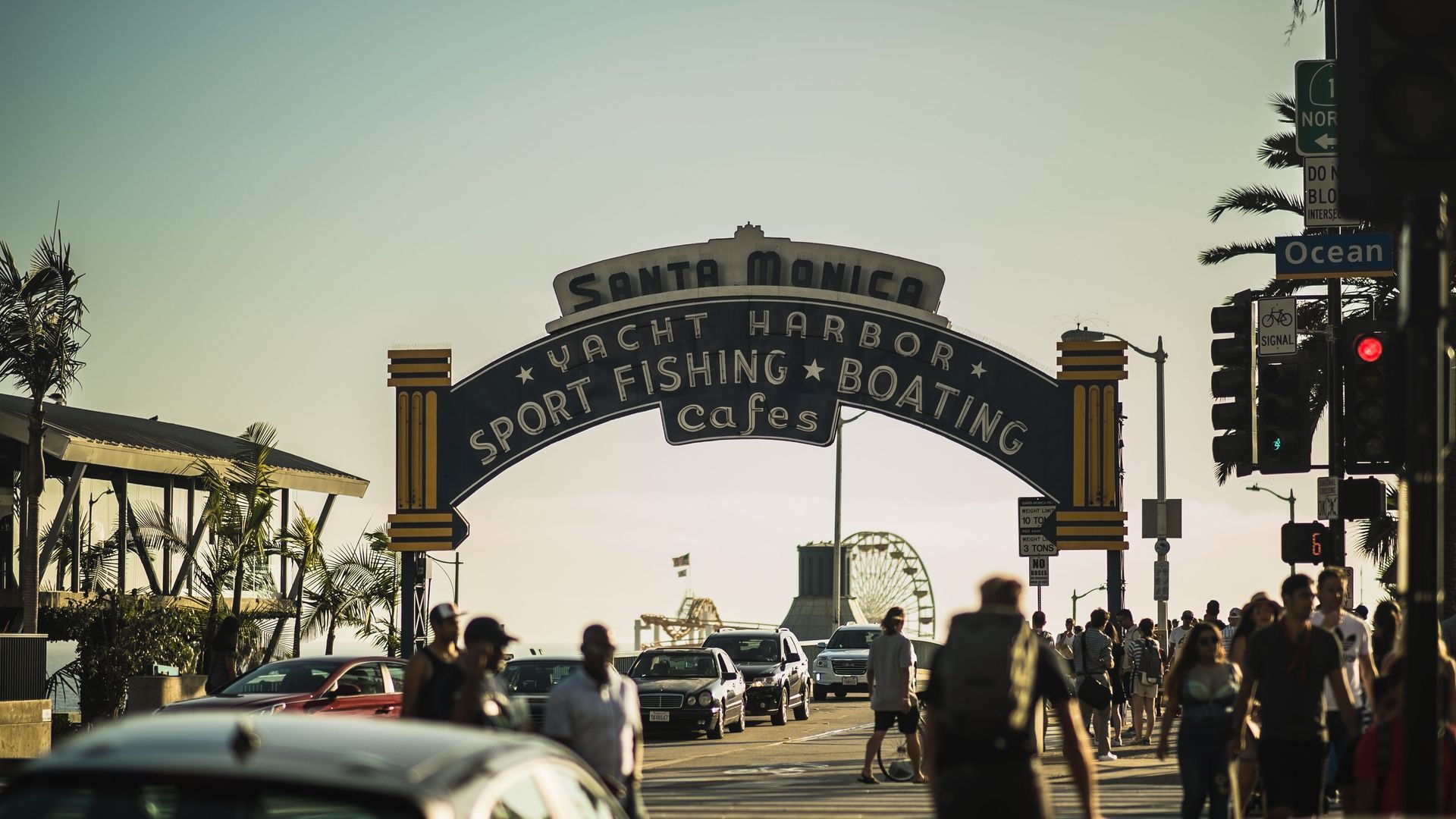 Santa Monica Pier arch over a sunny street with cars, pedestrians, and palm trees near the beach