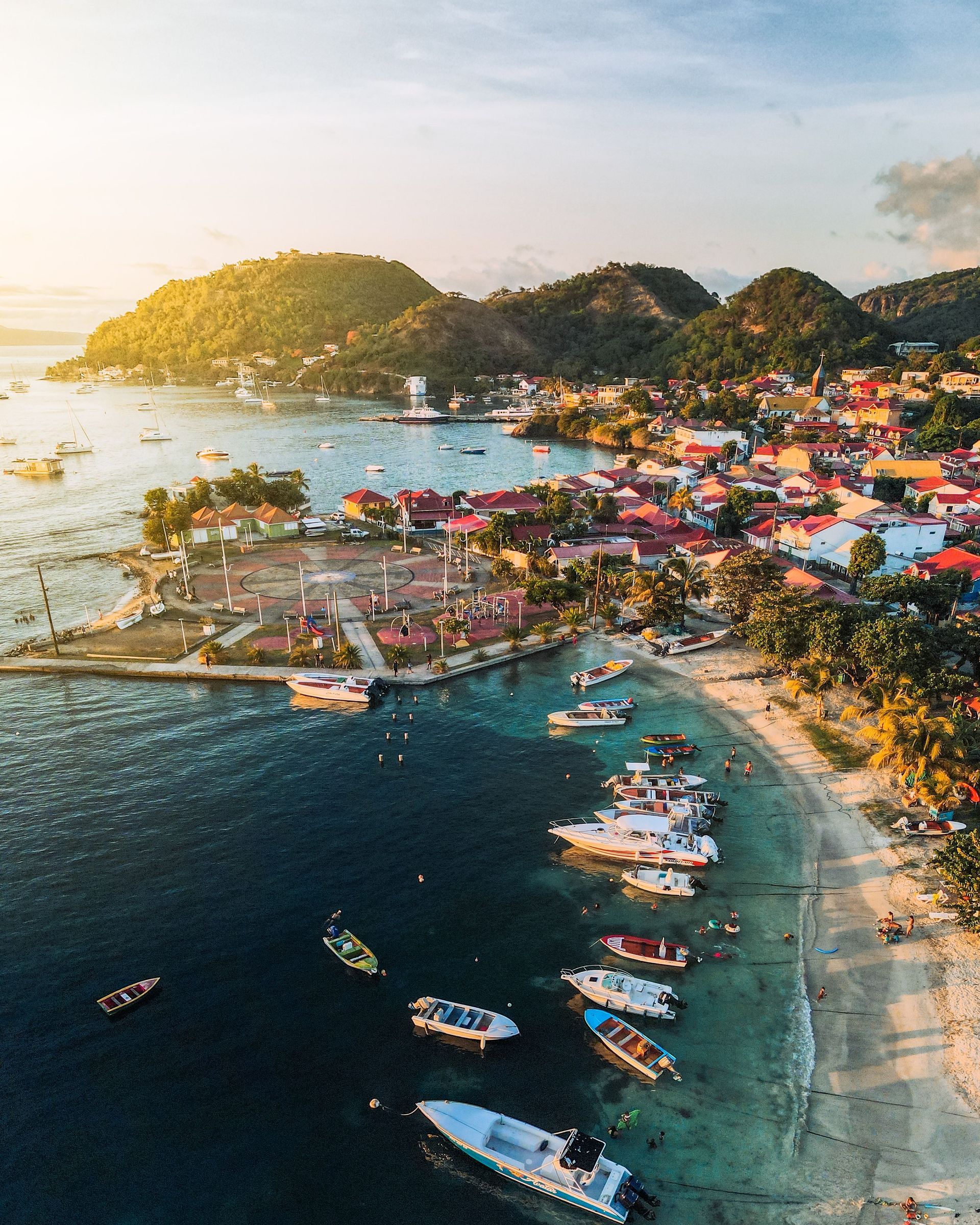 Aerial view of a colorful seaside harbor with boats, beach, and hillside buildings at sunset
