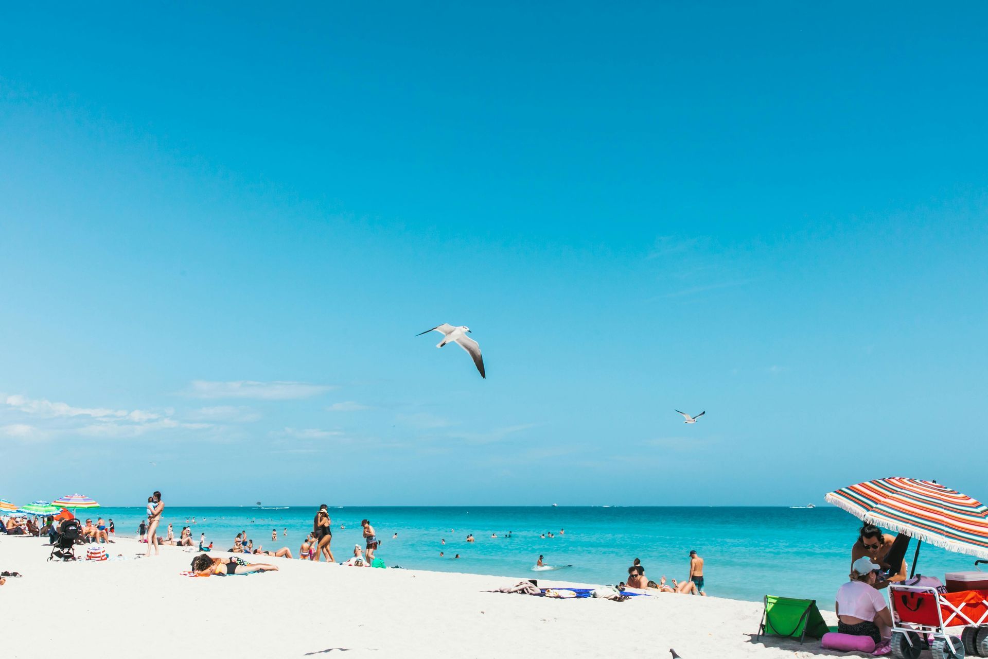 Sunny beach with turquoise water, people relaxing on the sand, and a striped umbrella under a blue sky.