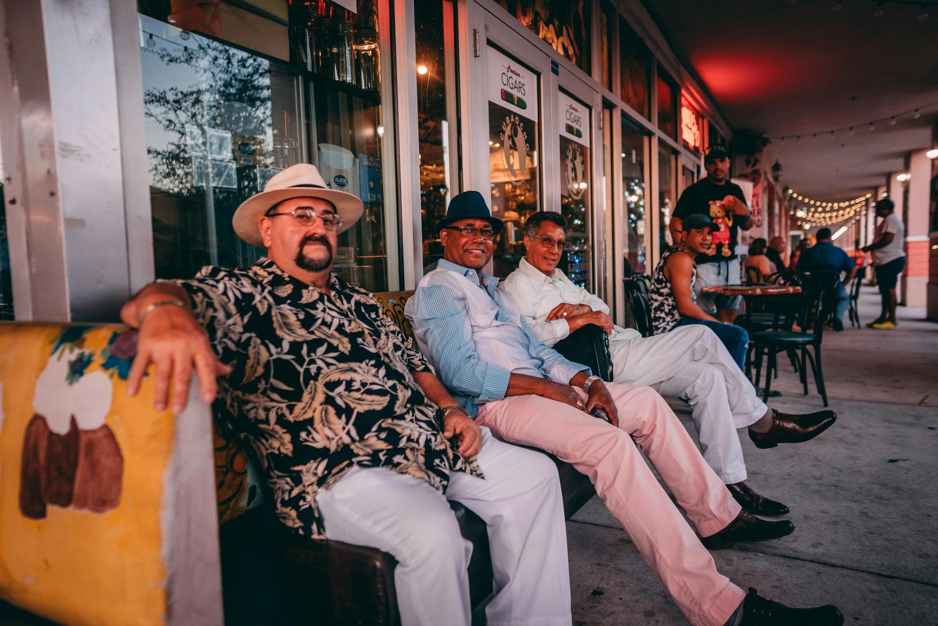 People seated on a sidewalk bench outside a storefront at dusk, with warm street lights and a relaxed atmosphere