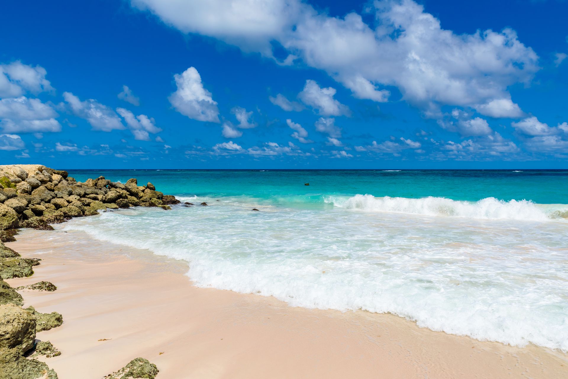 Sunny beach with turquoise waves, pale sand, and rocky shoreline under a blue sky with clouds