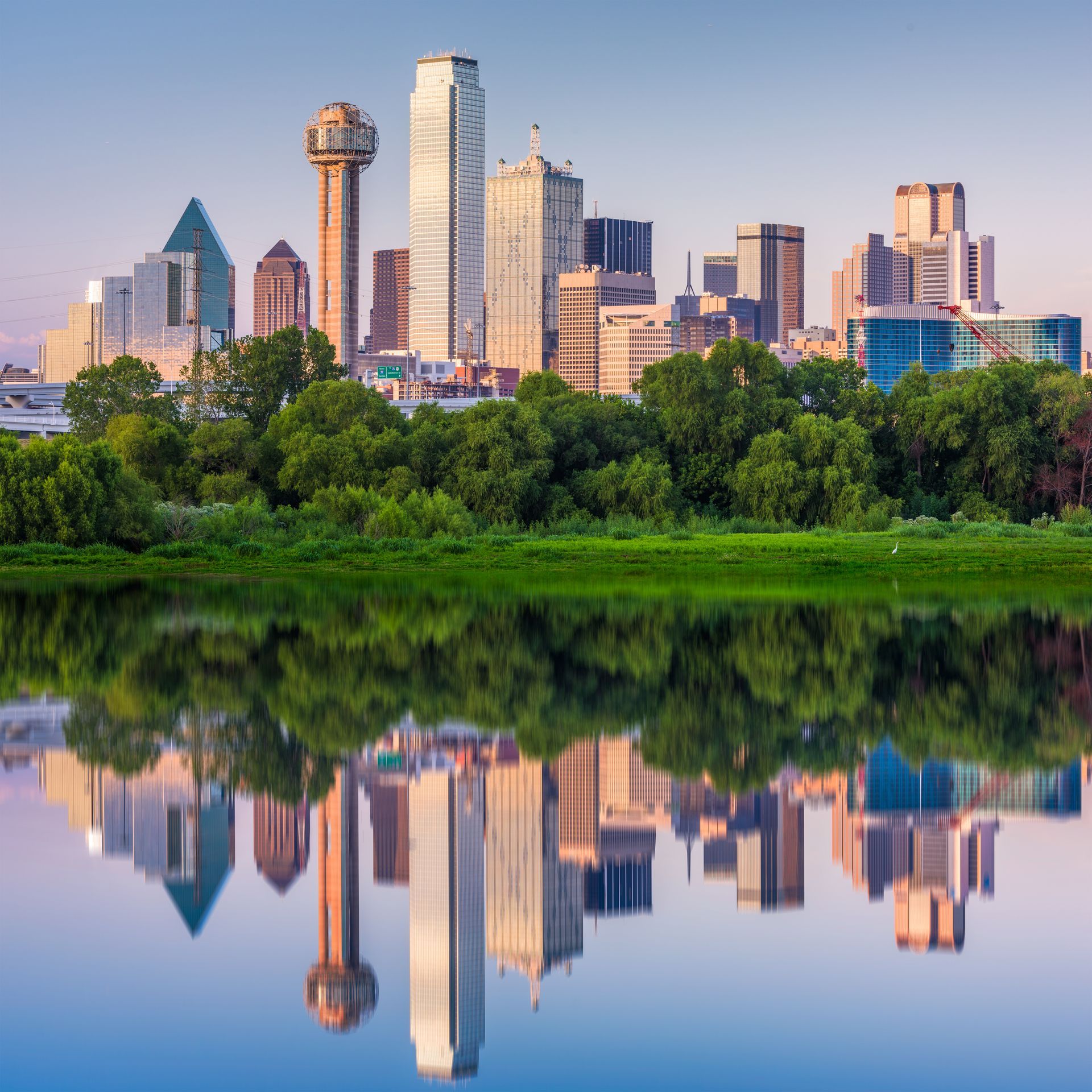 Downtown skyline reflected in a calm lake at sunrise, framed by green trees and pink sky