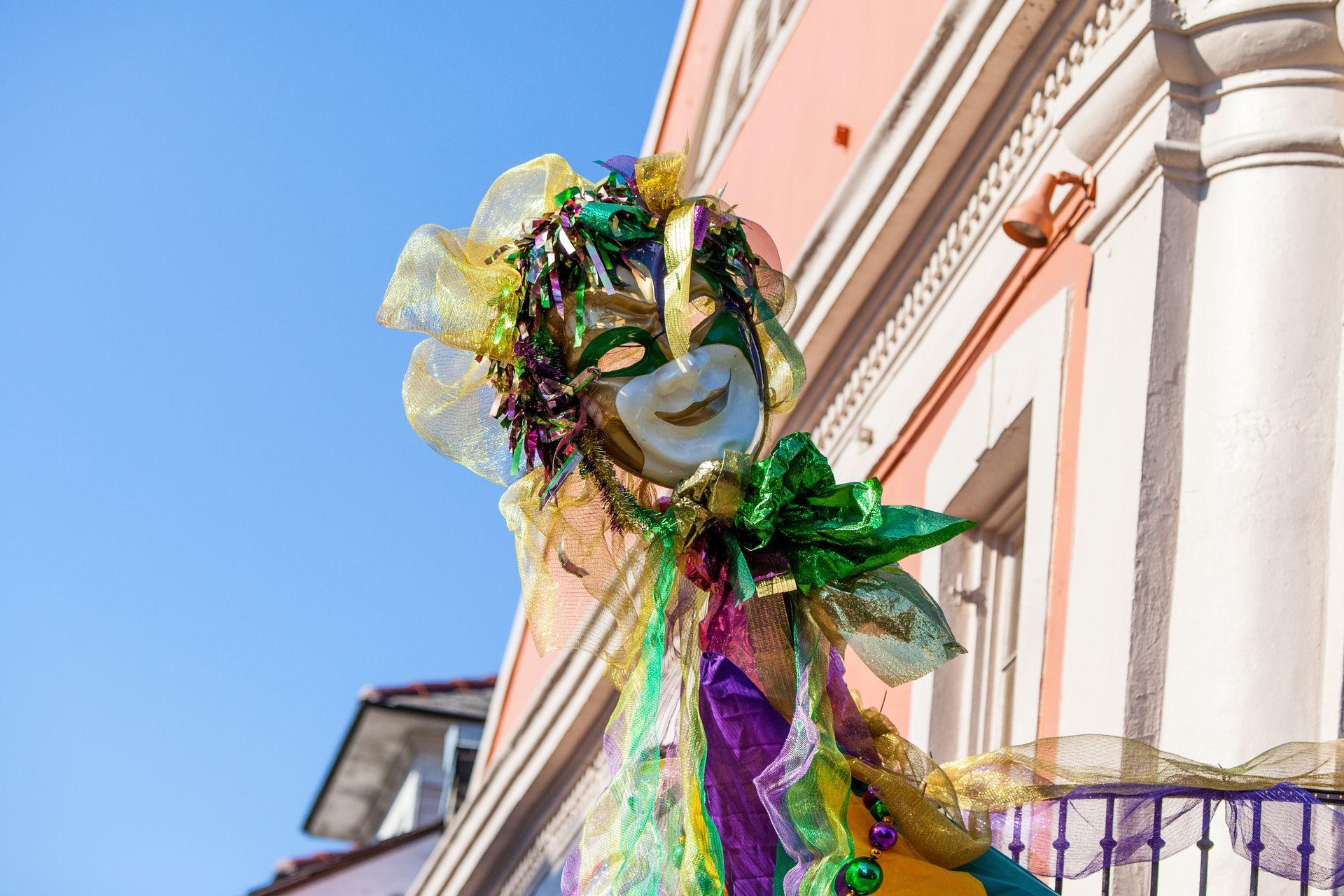 Colorful masked parade figure with green, purple, and gold ribbons outside a pastel building