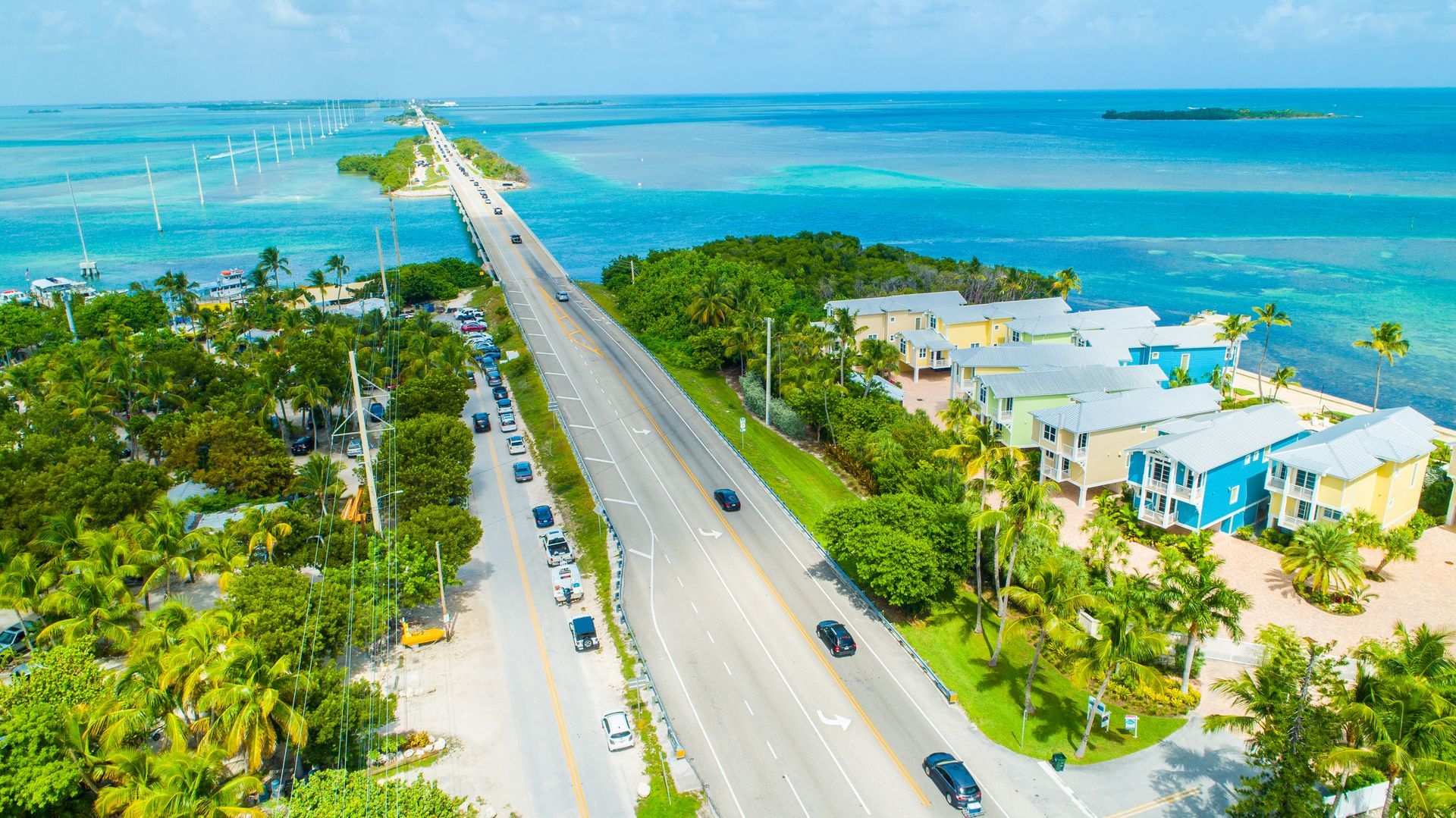 Aerial view of a coastal road over turquoise water beside green trees and pastel waterfront homes.