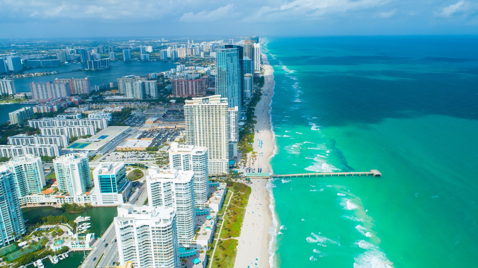 Aerial view of a coastal city skyline beside turquoise ocean and sandy beach.