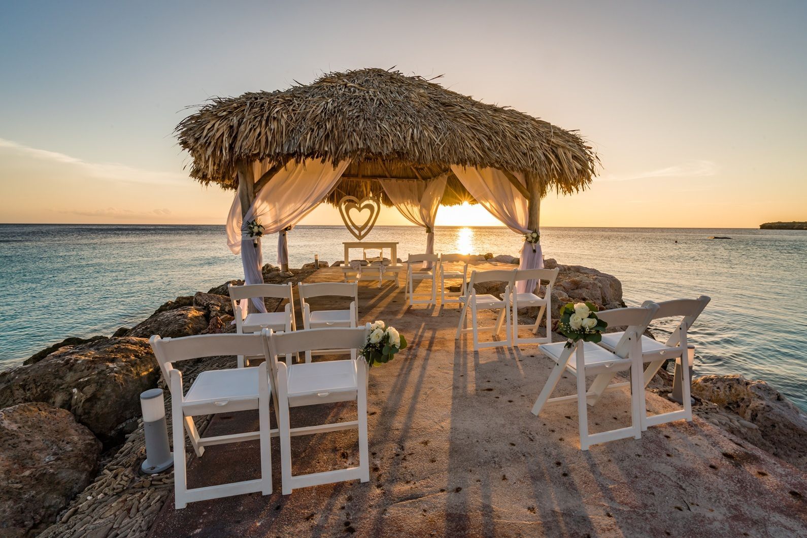 Beachside wedding arch with white chairs on a rocky shore at sunset
