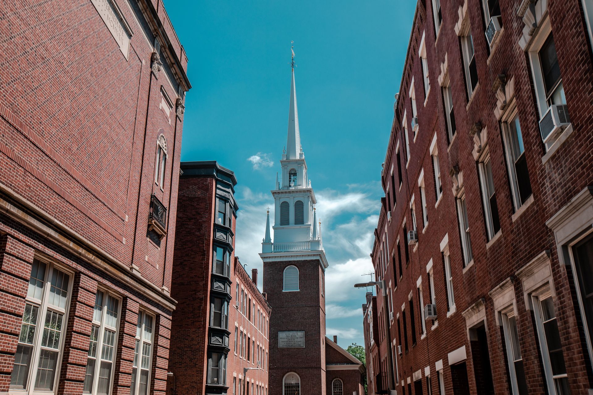 Brick buildings frame a church steeple against a blue sky in a narrow city street.