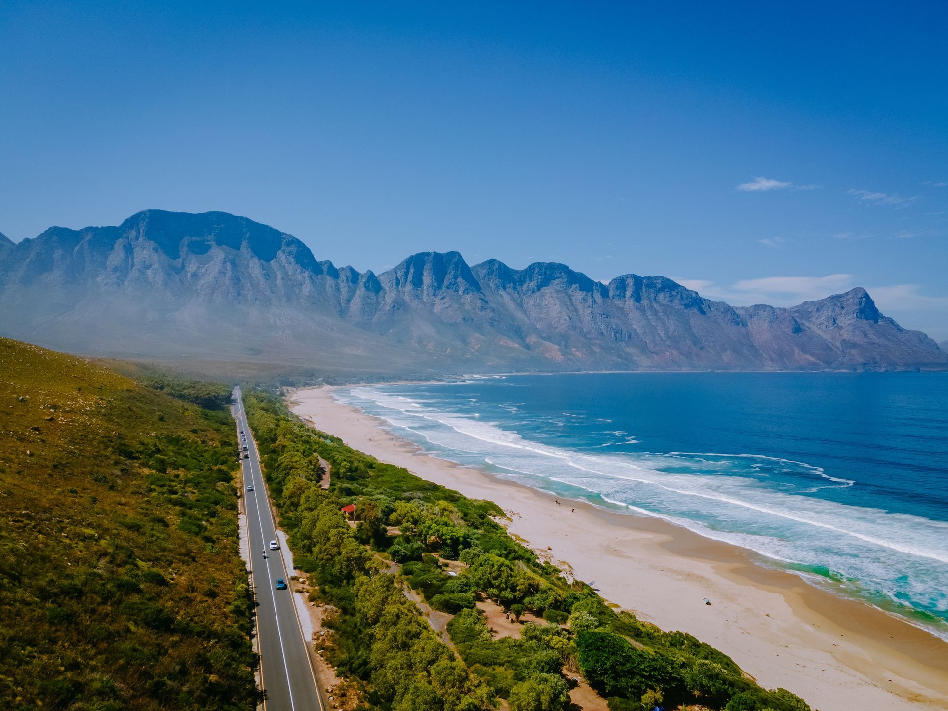 Aerial view of a coastal highway beside a sandy beach and blue ocean with mountains in the background
