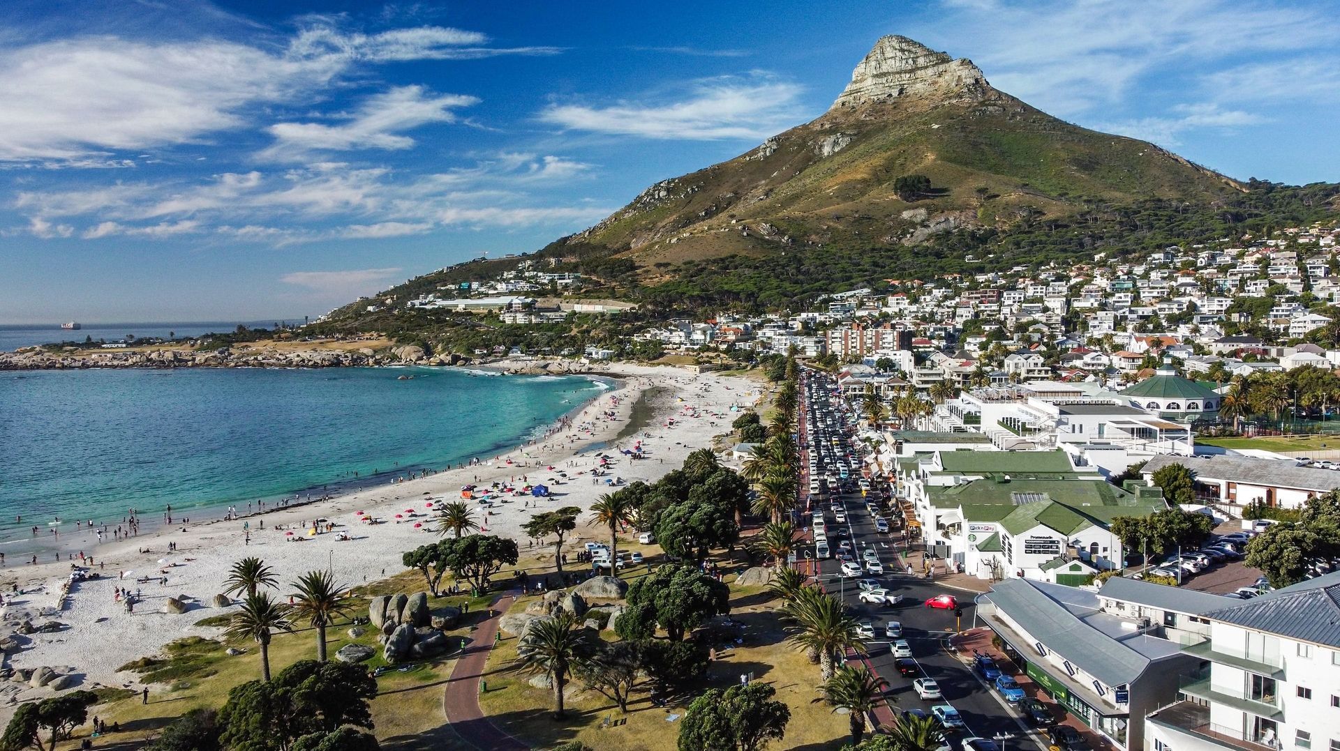 Coastal city with a turquoise bay, busy beachfront road, white buildings, and a mountain under a blue sky