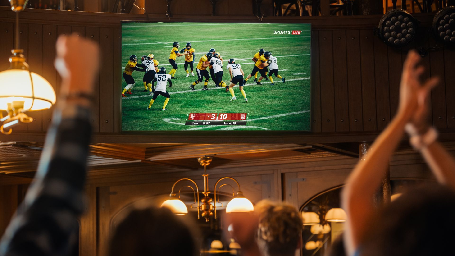 Crowd watching a football game on a wall-mounted TV in a warm, dimly lit bar
