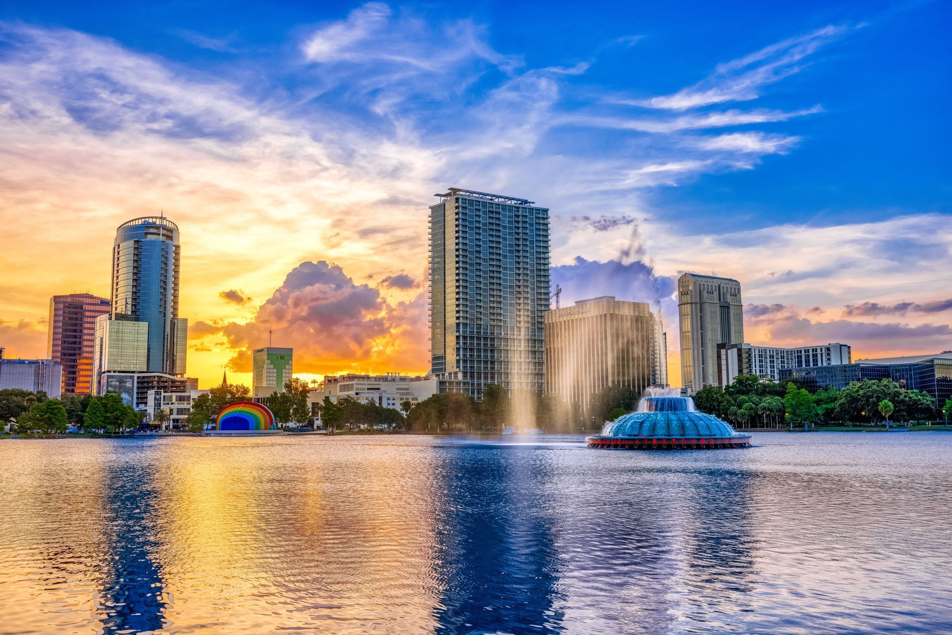 Sunset skyline with tall buildings, a lake, and a fountain spraying in the foreground