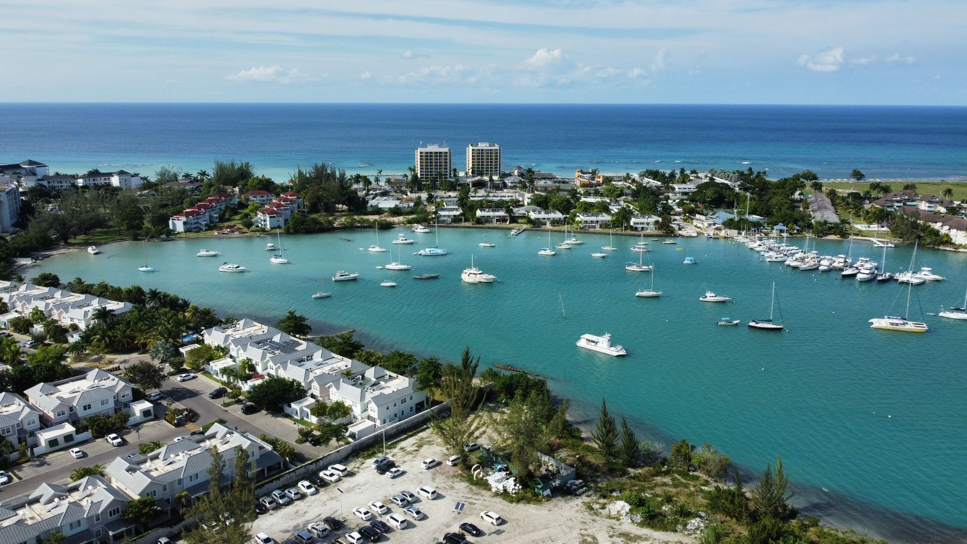Aerial view of a turquoise harbor with anchored boats, palm-lined shores, and ocean beyond