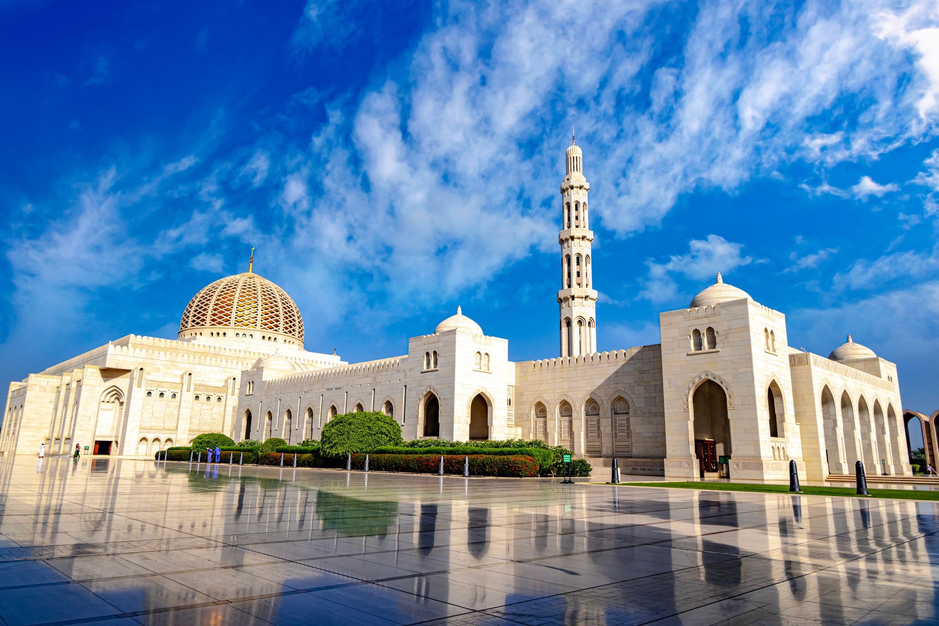 White mosque with golden dome and minaret reflected in wet pavement under a blue sky
