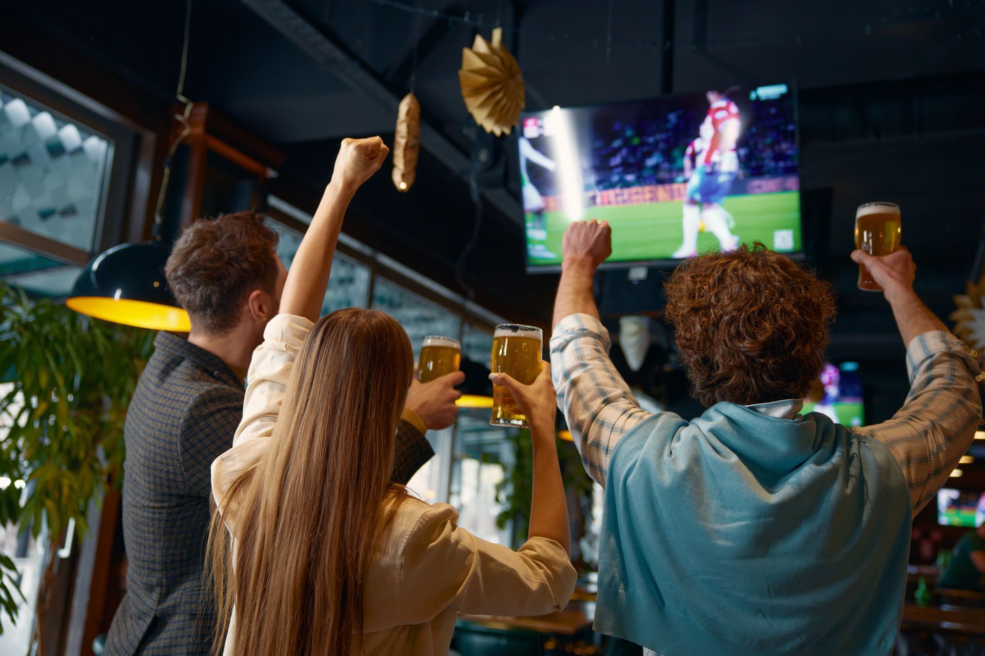 People cheering with drinks while watching a soccer match on a large TV in a bar