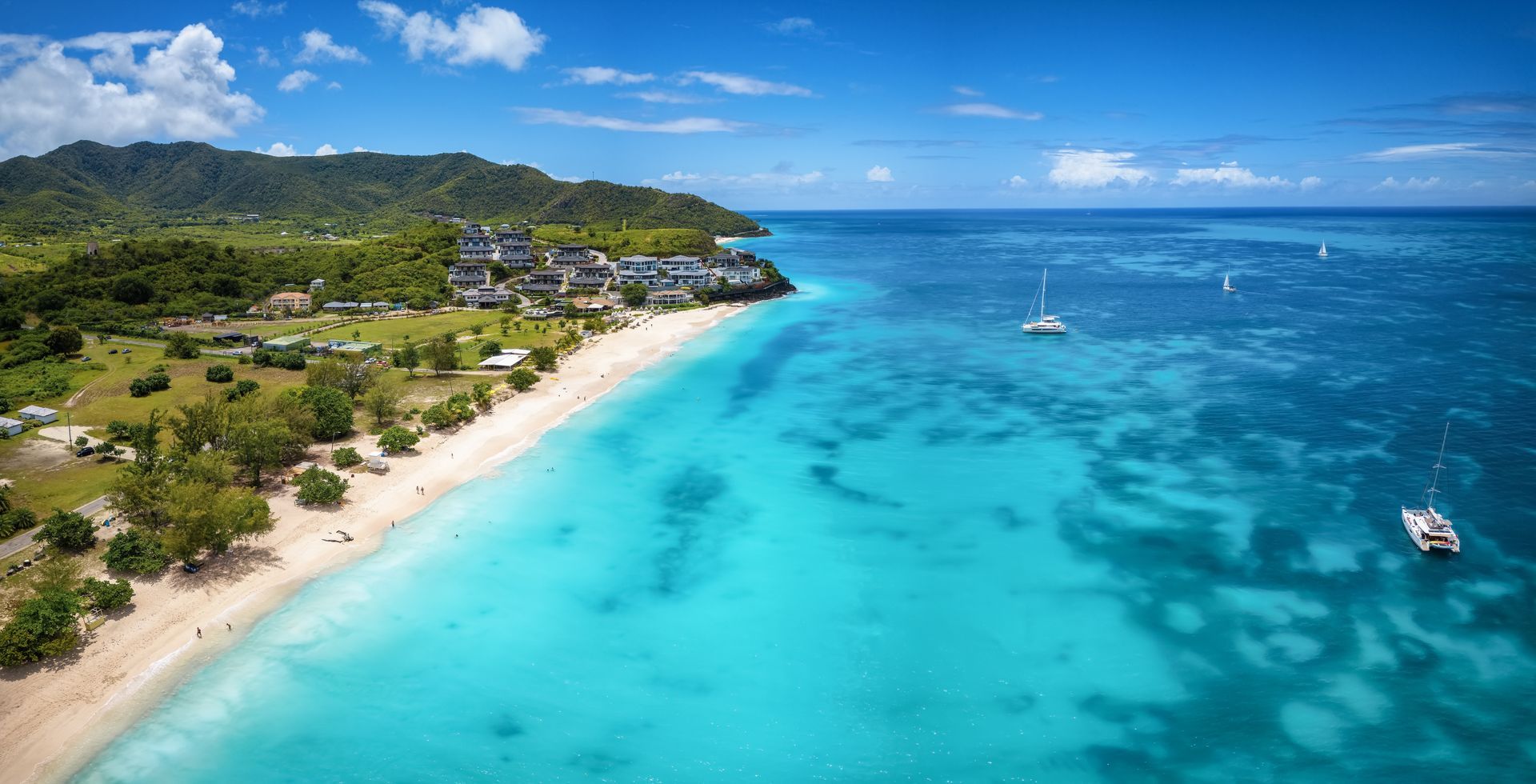 Aerial view of a turquoise tropical bay, sandy beach, green hills, and small boats under a blue sky