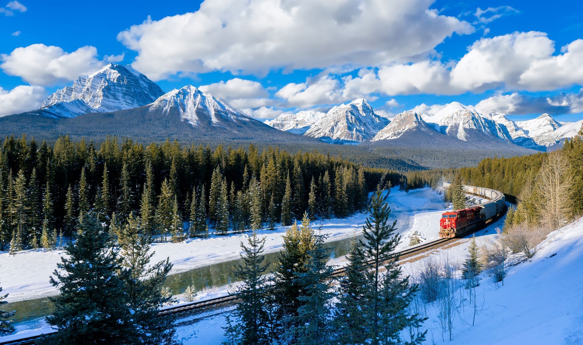 Snowy mountain landscape with pine forest and a red train curving through the valley under cloudy skies