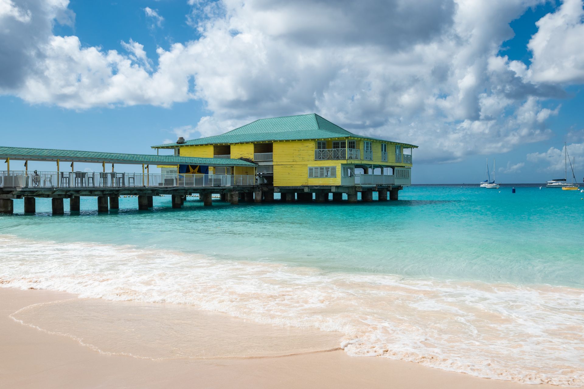 Tropical beach with a yellow overwater building and turquoise water under a partly cloudy sky