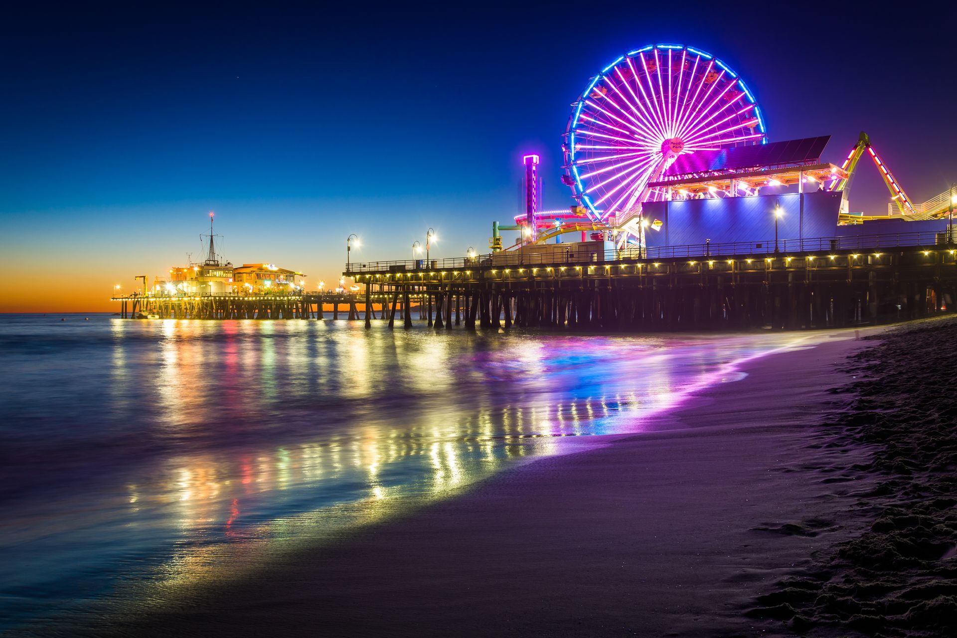 Colourful Ferris wheel and lit Santa Monica pier at night, reflecting on the calm ocean shoreline
