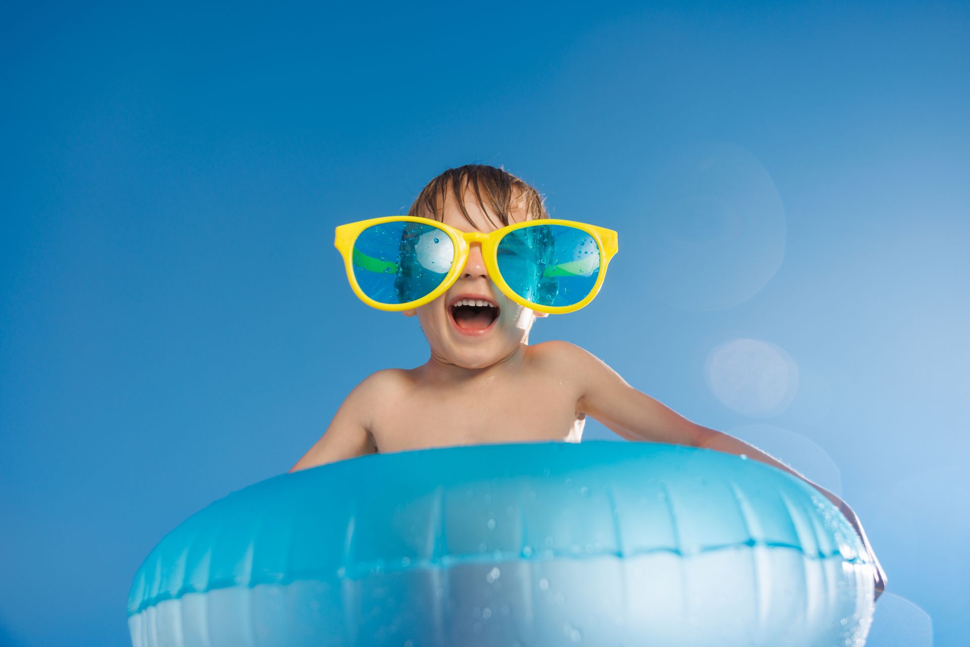 Child in yellow sunglasses smiling in a blue inflatable pool against a blue sky background