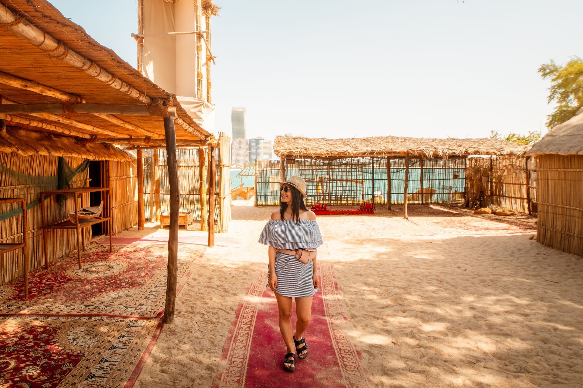 Person walking on a red carpet in a bright outdoor courtyard with rustic buildings and shade structures.