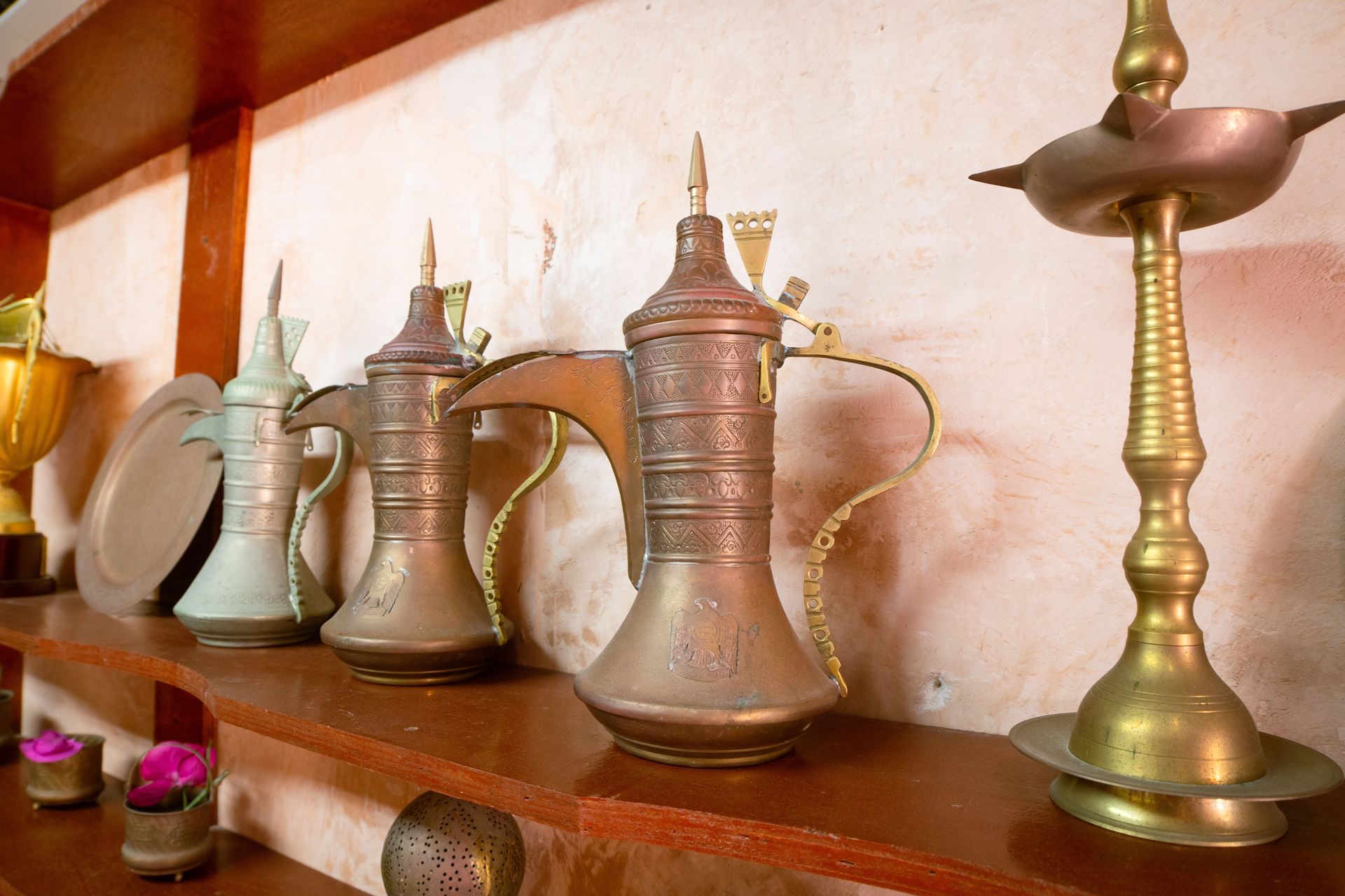 Row of ornate metal coffee pots and a brass lamp on a wooden shelf against a white wall