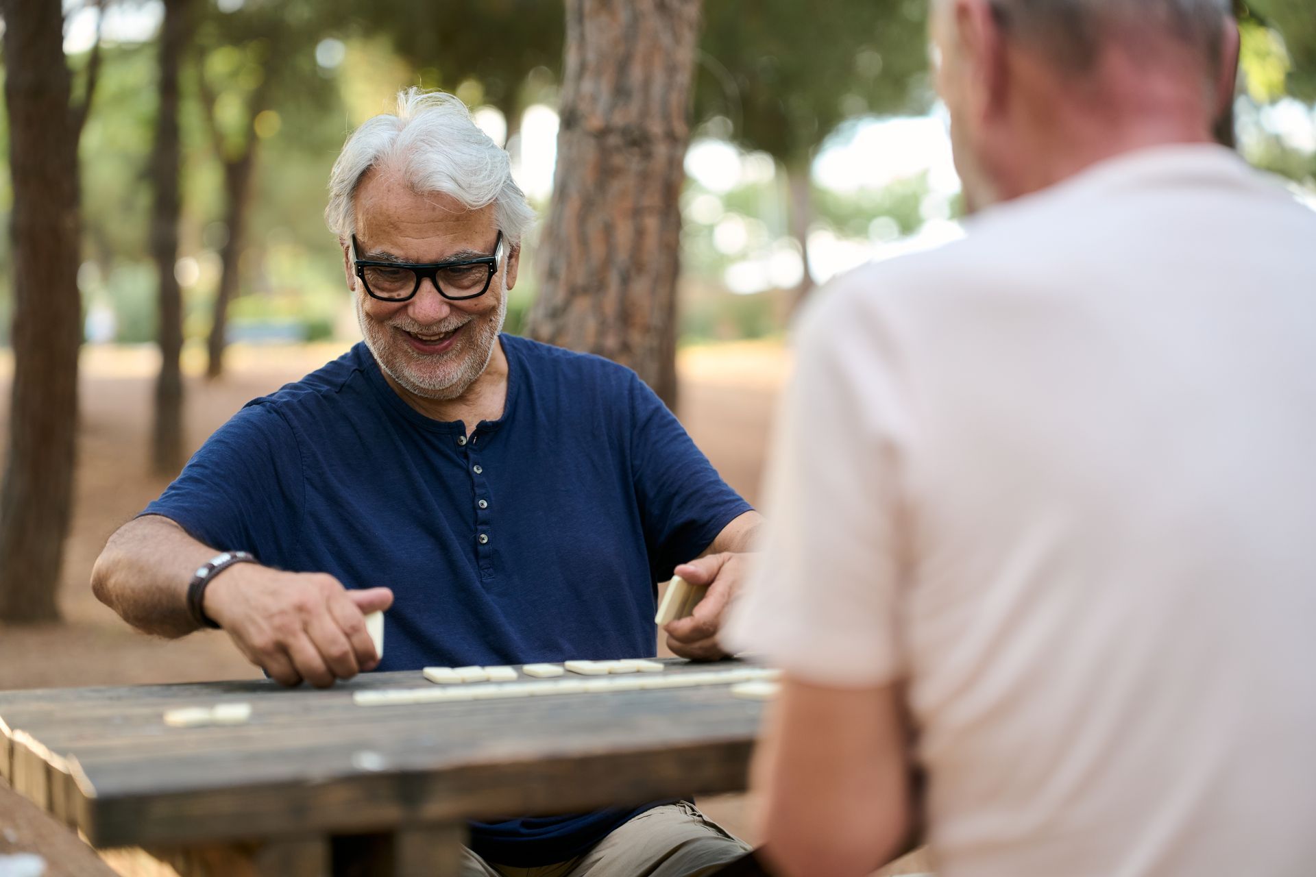 Two people talking at a wooden picnic table outdoors, one smiling and gesturing with his hand.