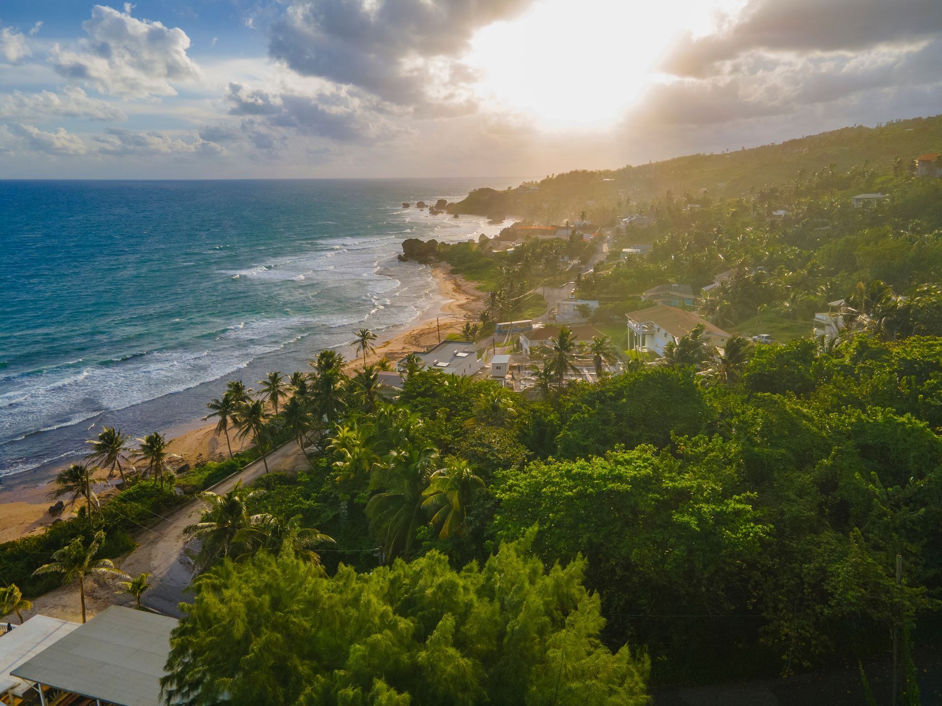 Sunlit tropical coastline with blue ocean, sandy beach, palm trees, and green hills under cloudy sky