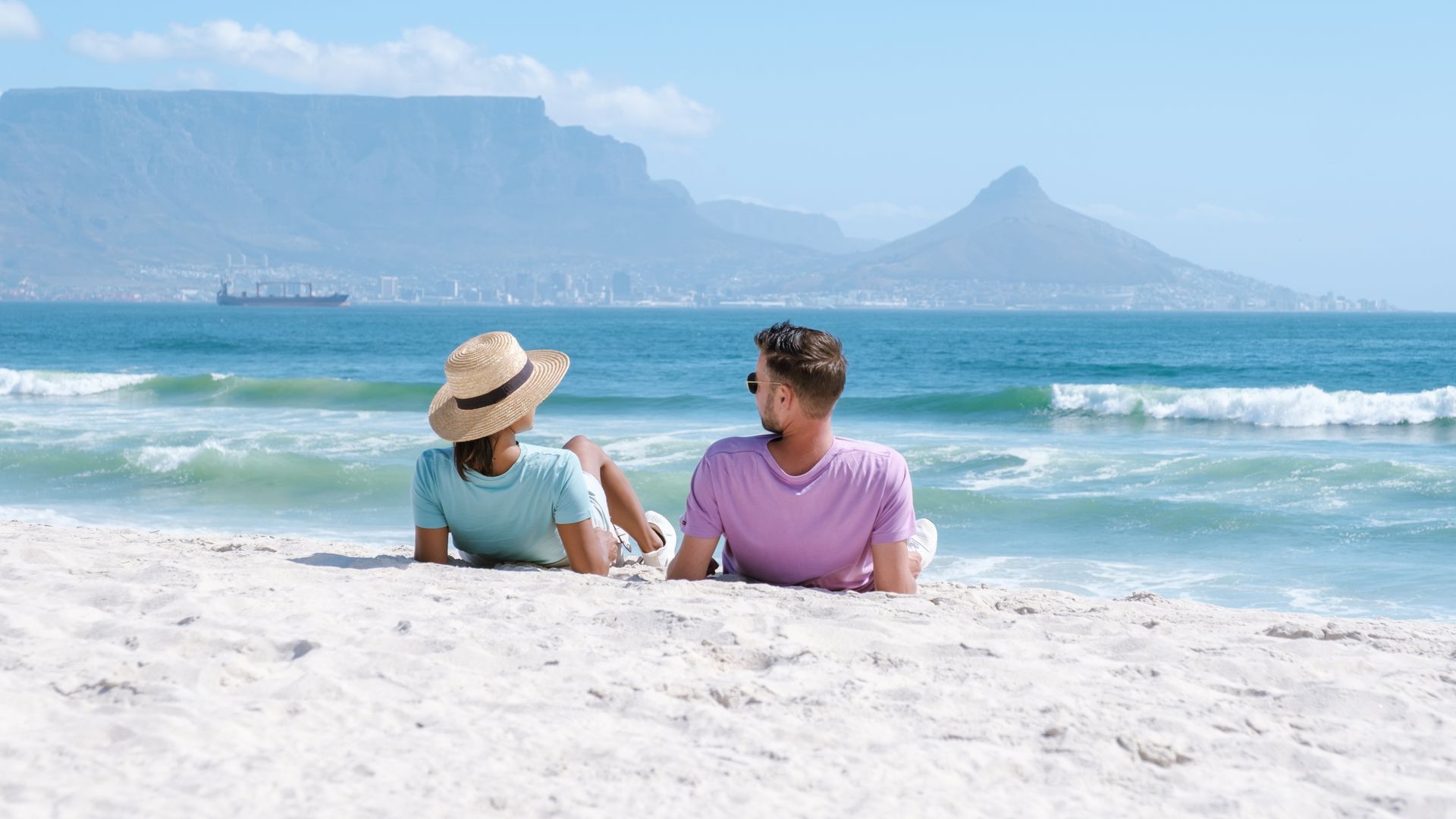 Two people sit on a sandy beach facing the ocean and distant mountains under a clear blue sky.