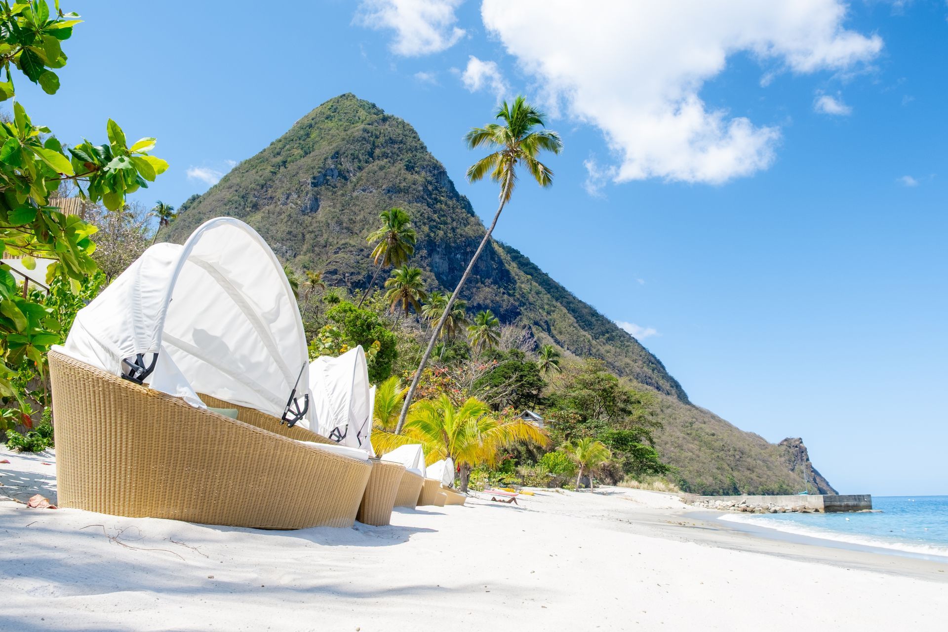 Lounge chair on a bright white-sand beach with palm trees, turquoise water, and a rocky green mountain under blue sky