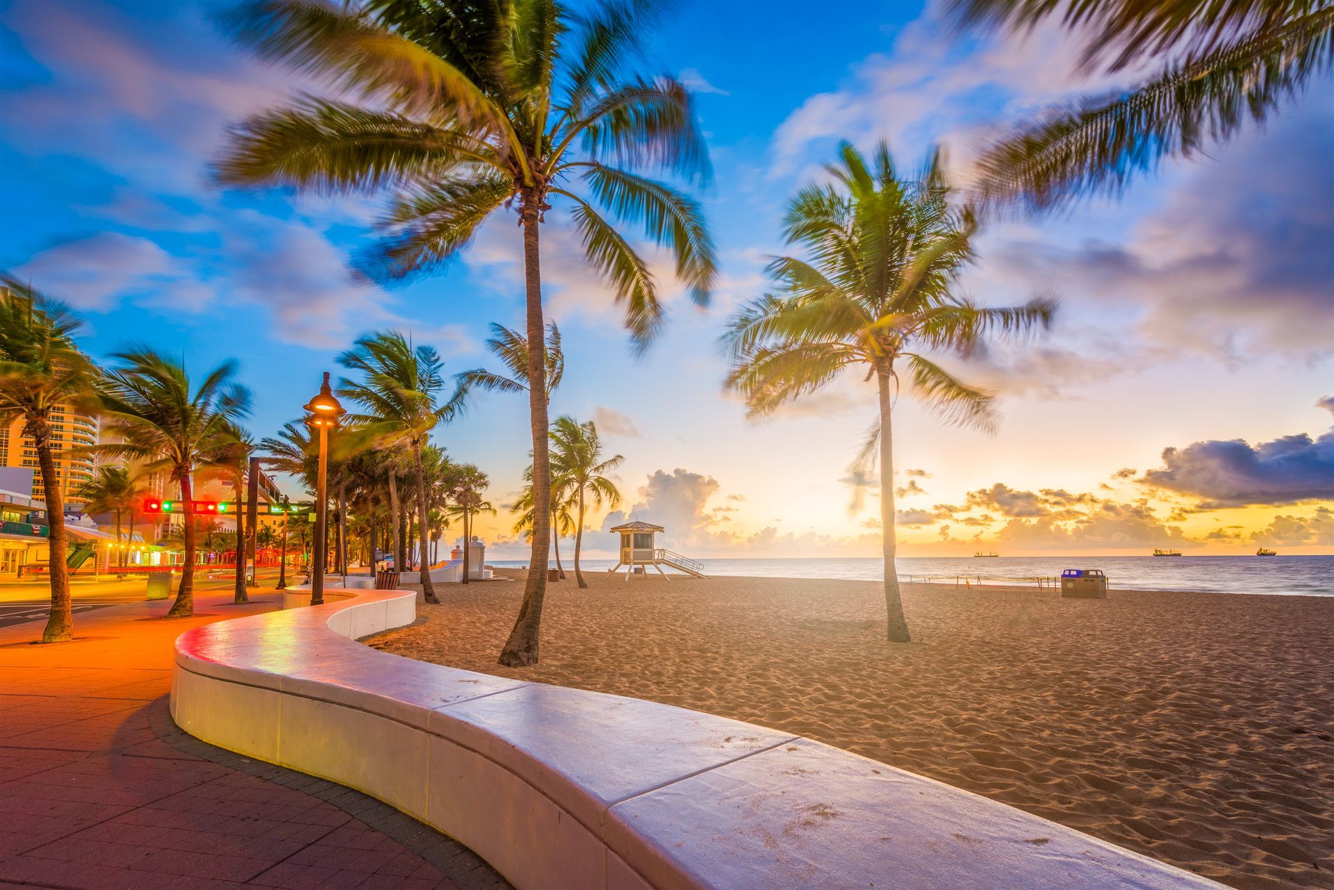 Sunset beach with palm trees, curved promenade, and calm ocean under a colorful sky