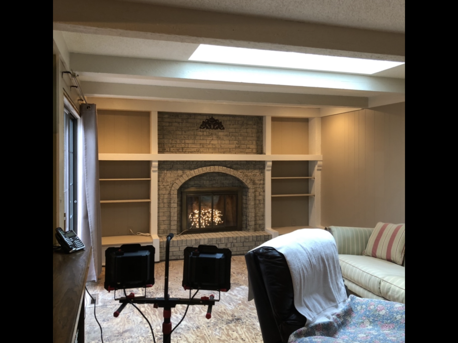 Living room with fireplace, built-in shelves, and a skylight. A couch and chair are in the foreground, and there are two studio lights.