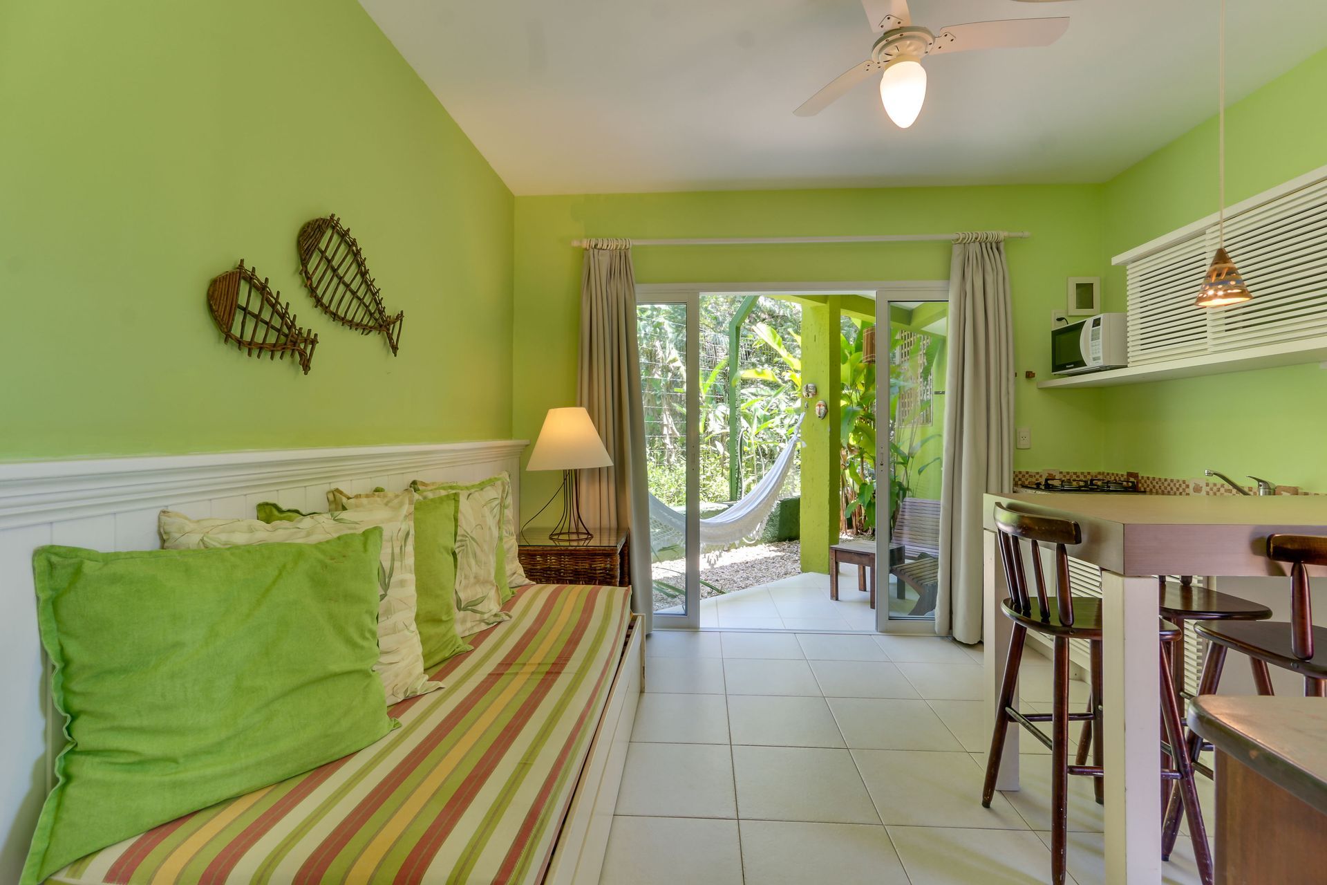 Quarto tropical luminoso com paredes verdes, cama listrada, portas abertas para um pátio e uma kitchenette.