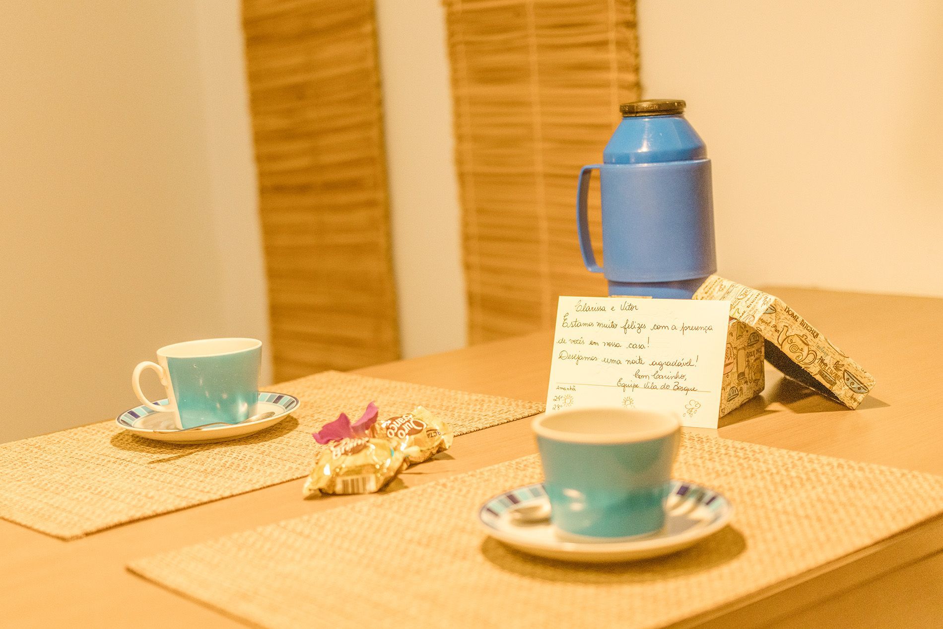 Conjunto de chá sobre uma mesa de madeira com xícaras azuis, um bule e um bilhete escrito à mão ao lado de pequenos petiscos.