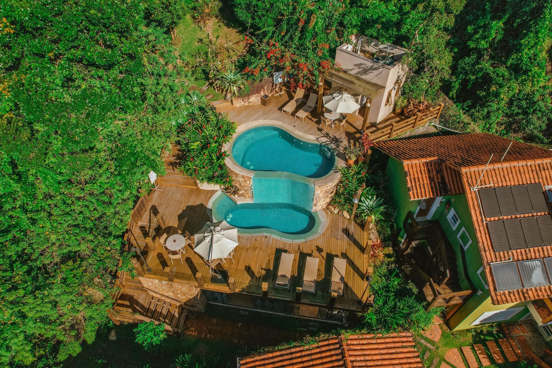 Vista aérea da piscina de um resort rodeada por vegetação exuberante e edifícios com telhados de telha vermelha.