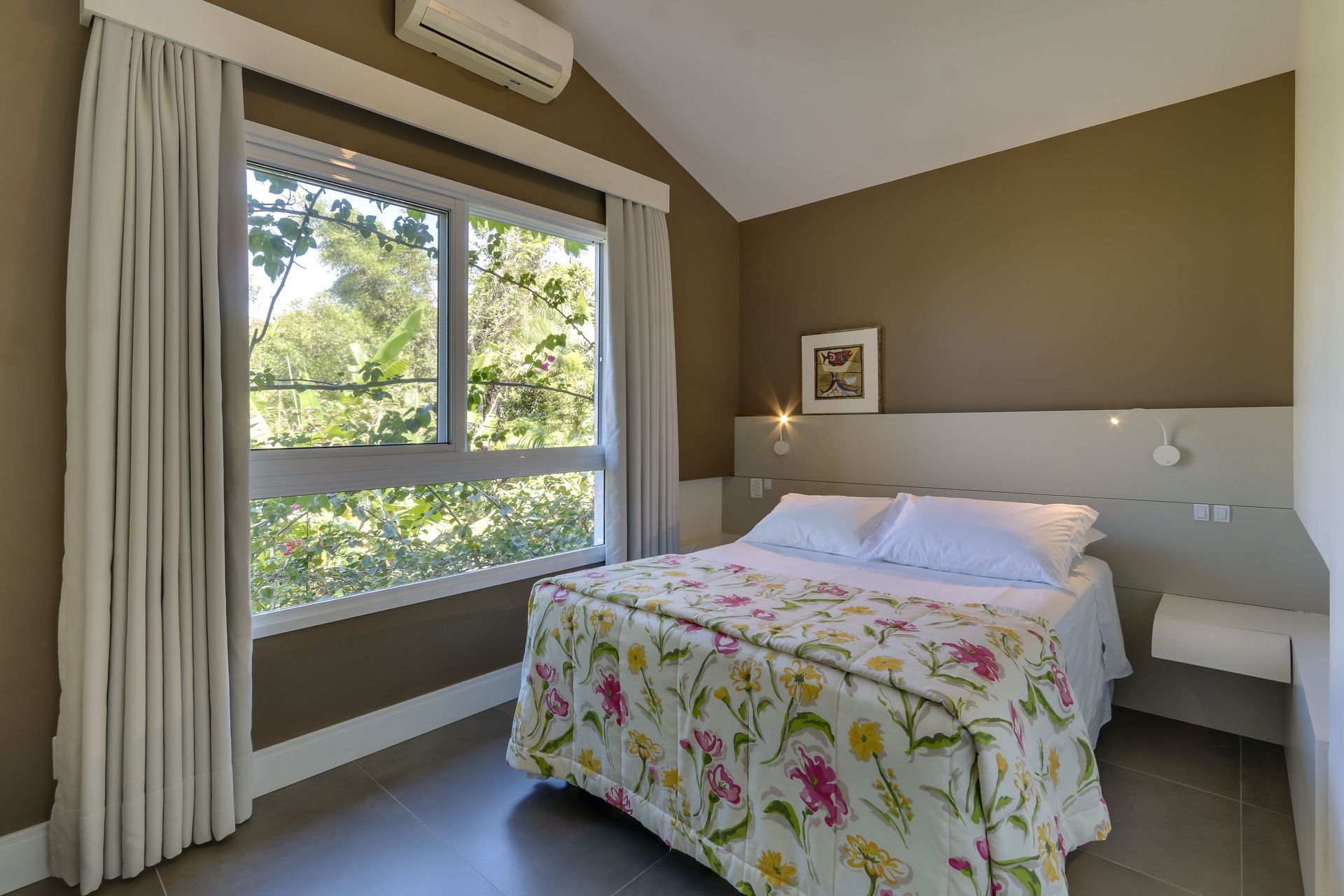 Quarto luminoso com uma grande janela, paredes bege e uma colcha floral sob uma suave luz natural.
