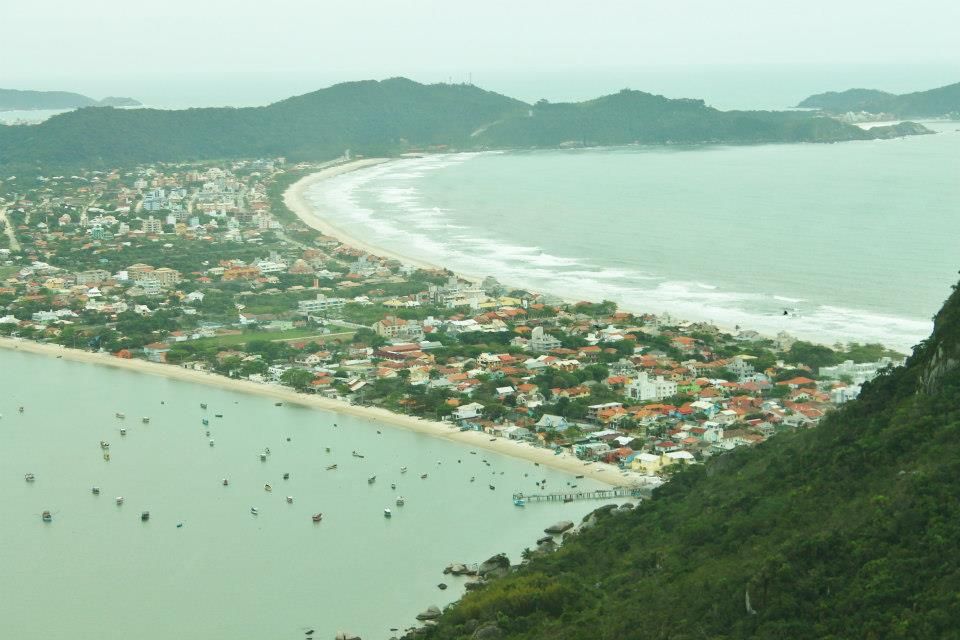 Vista aérea de uma cidade costeira que se estende ao longo de uma baía com colinas verdejantes e águas calmas sob um céu enevoado.