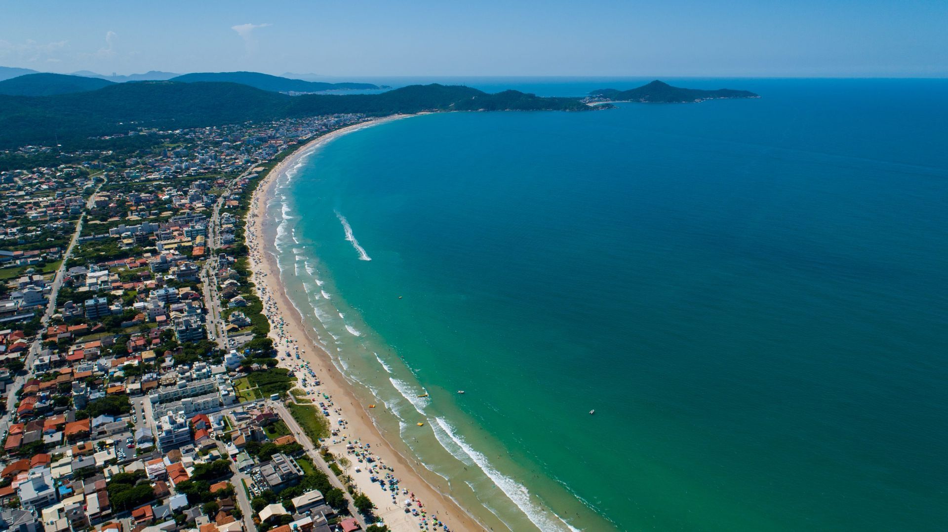 Vista aérea de uma cidade costeira que se estende ao longo de uma praia de areia, com águas turquesa e montanhas ao longe.