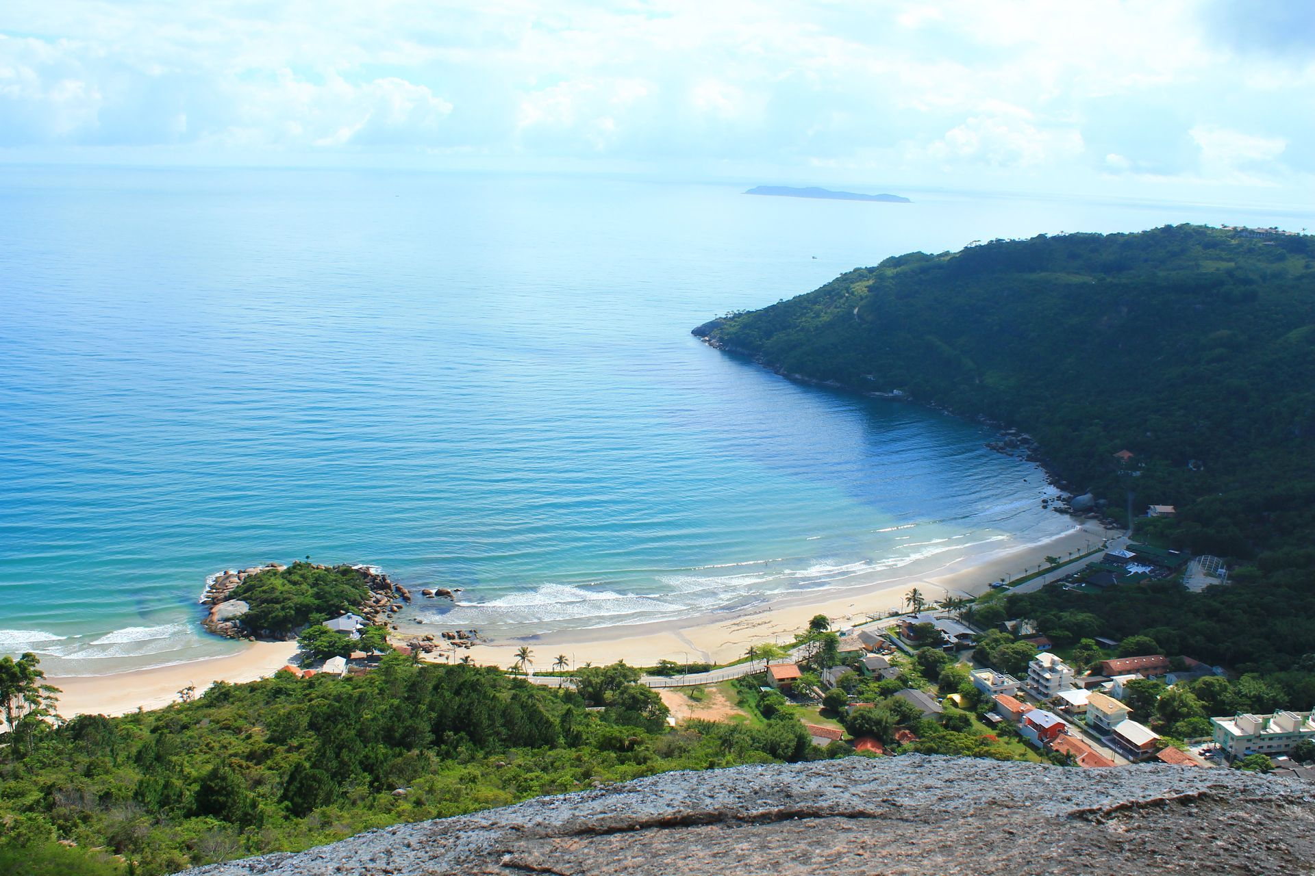 Baía costeira com água turquesa, praia de areia, encosta verdejante e pequena vila sob um céu azul.