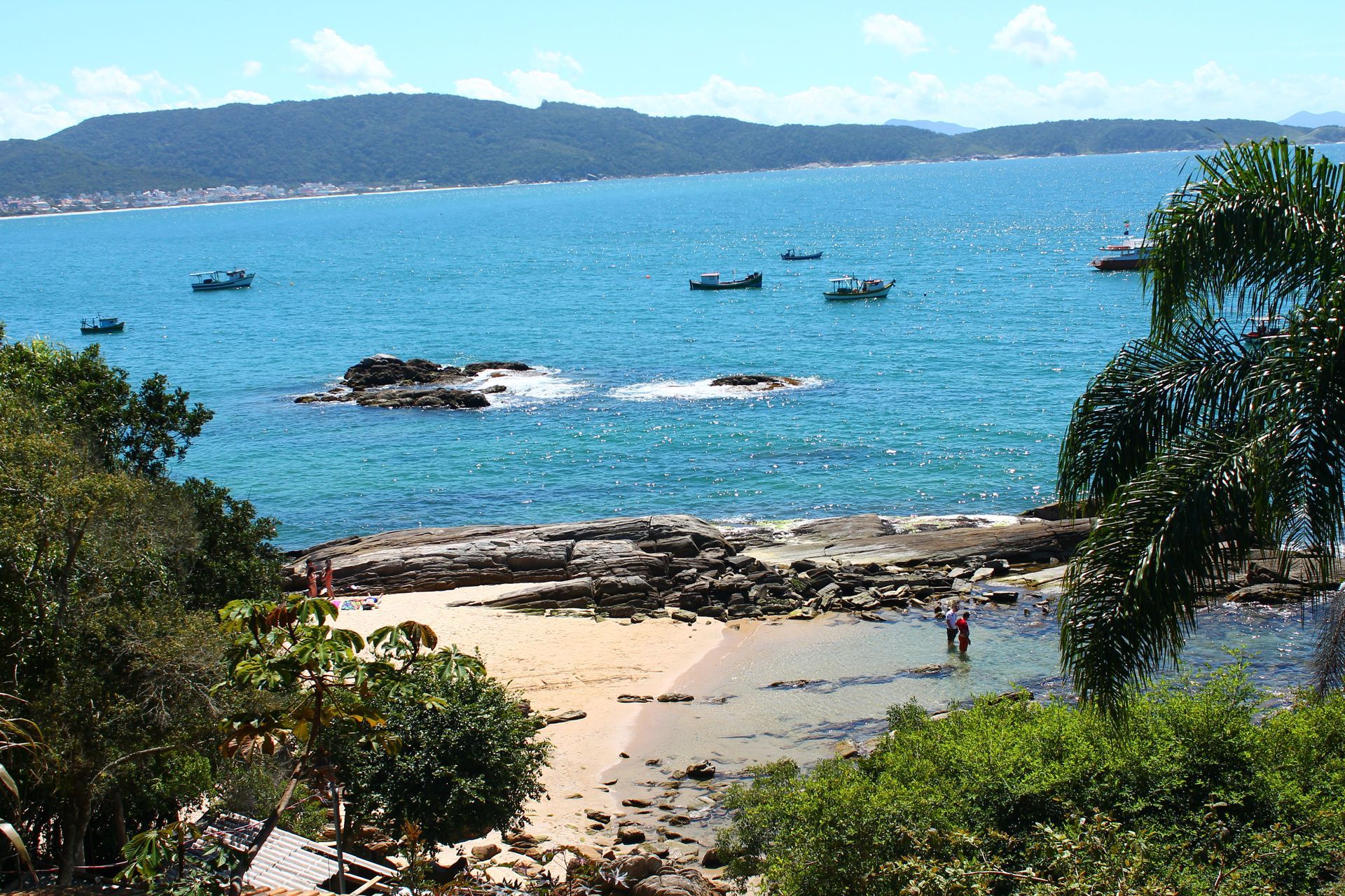 Praia tropical ensolarada com água turquesa, costa rochosa, barcos ao longe e folhagem verdejante.