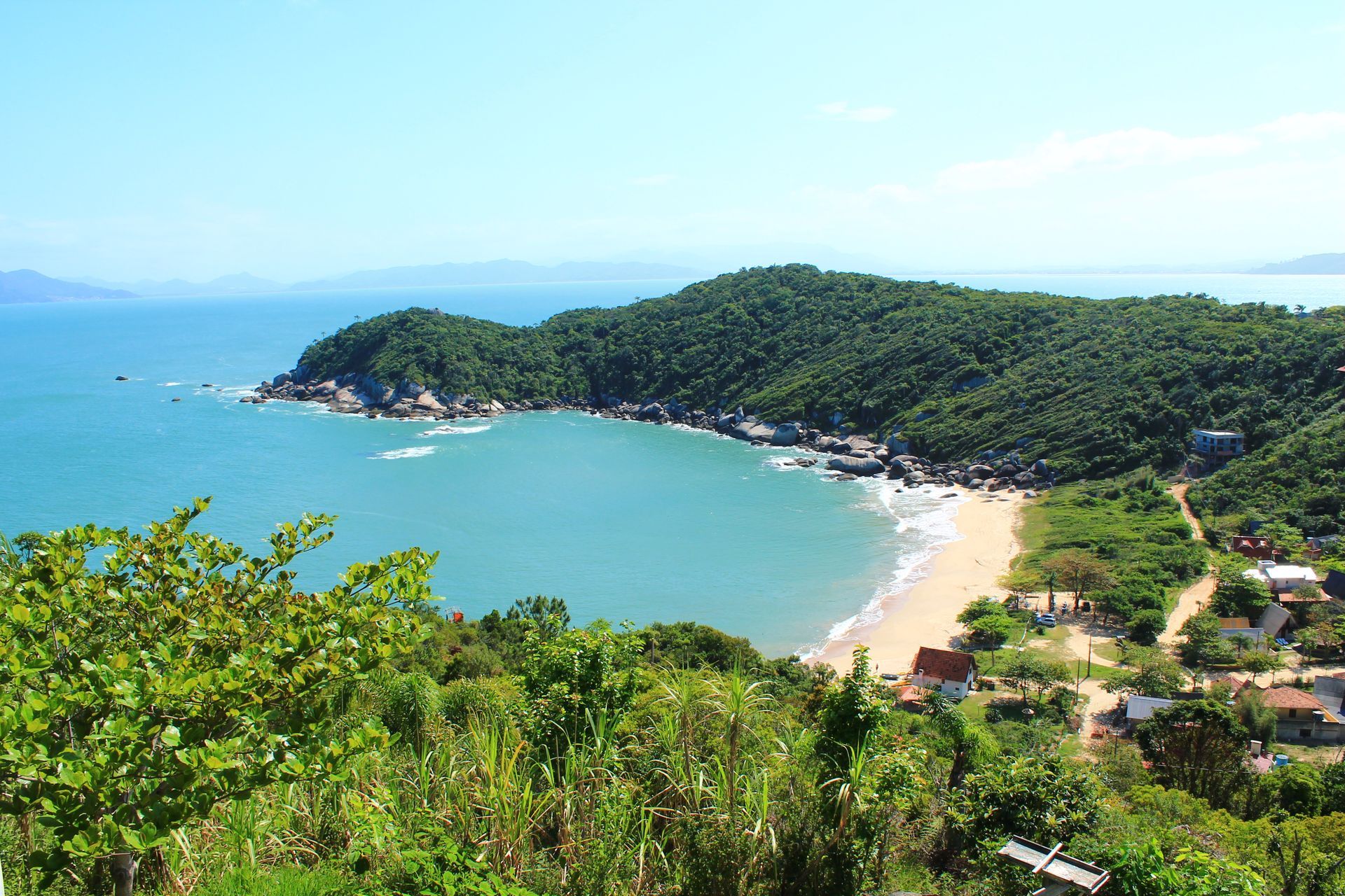 Baía tropical com água turquesa, praia de areia e colinas verdejantes sob um céu azul límpido.