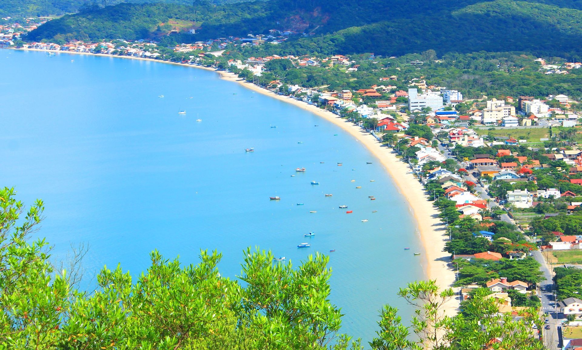 Vista aérea de uma praia de areia curva com água turquesa, casas à beira-mar e colinas verdes.