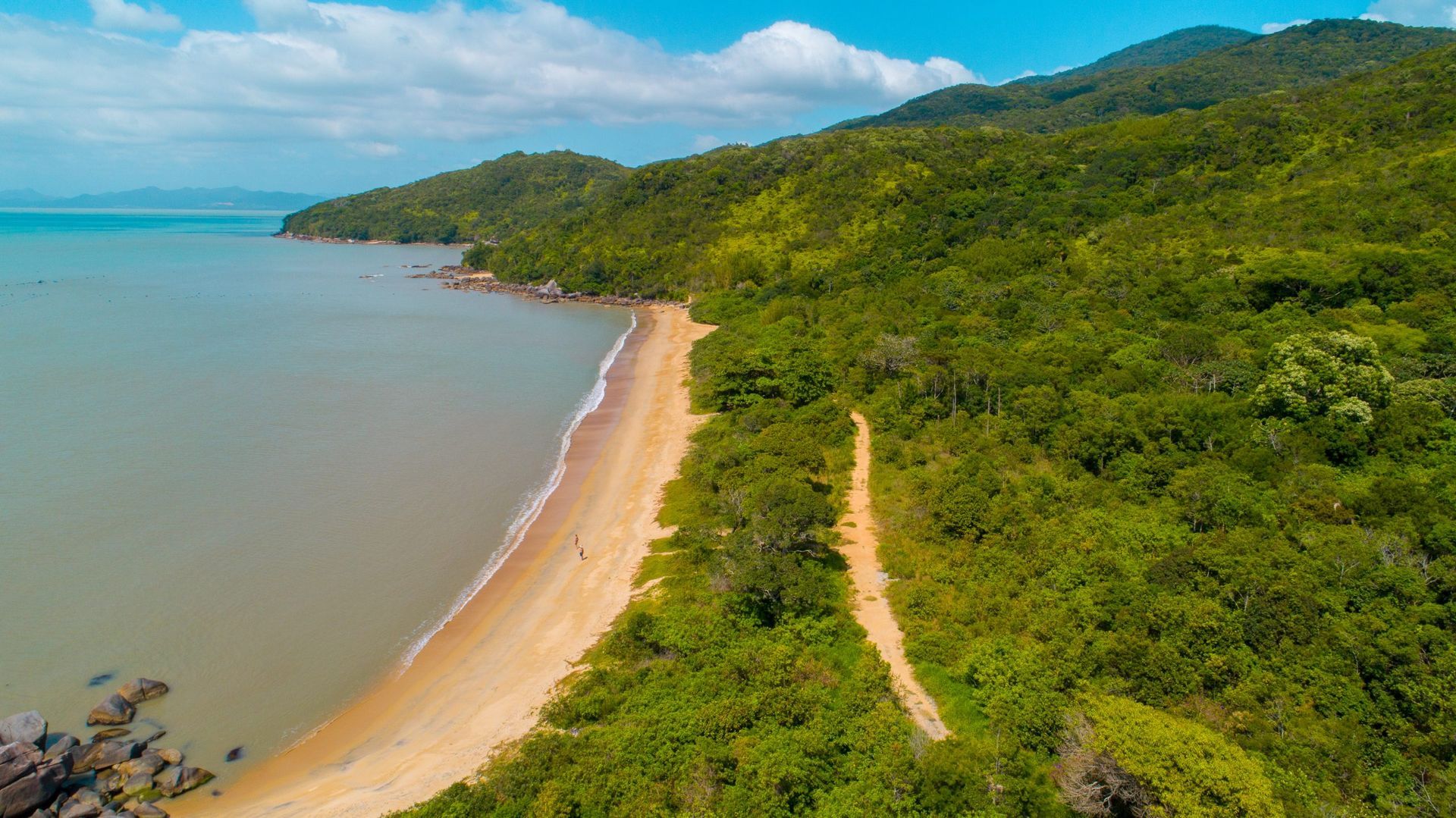 Vista aérea de uma praia tropical com água turquesa, faixa de areia e colinas verdejantes sob um céu azul.