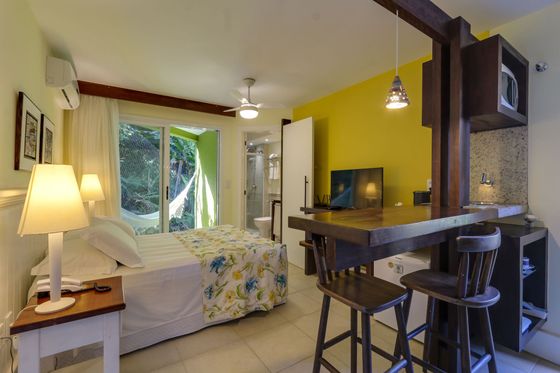 Estúdio aconchegante com cama, escrivaninha, banquetas e minicozinha sob paredes verdes.